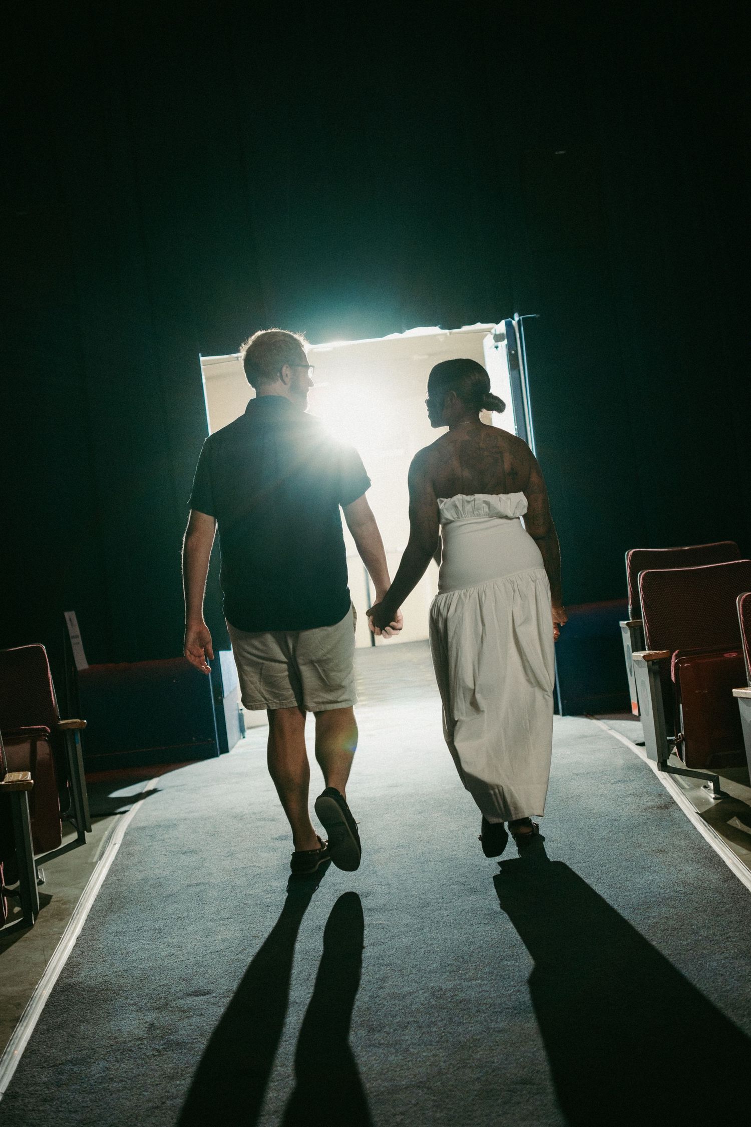 Couple holding hands walking toward bright doorway in dark theater aisle, silhouetted against glowing light.
