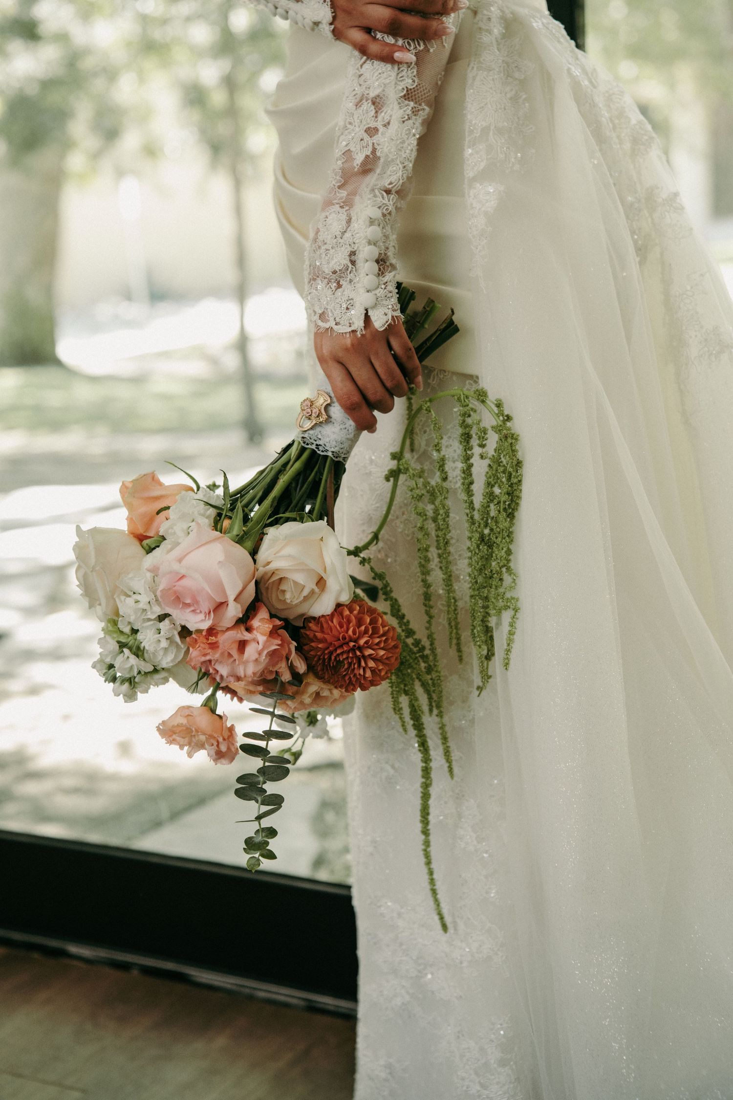 Bride in lace wedding dress holding elegant bouquet of pink roses, dahlias, and greenery.