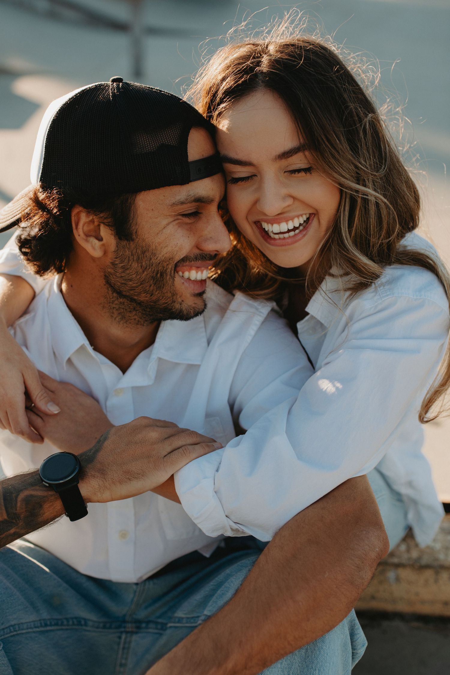 A laughing couple in white shirts embrace outdoors, the man wearing a black cap and watch, sharing a joyful romantic moment.