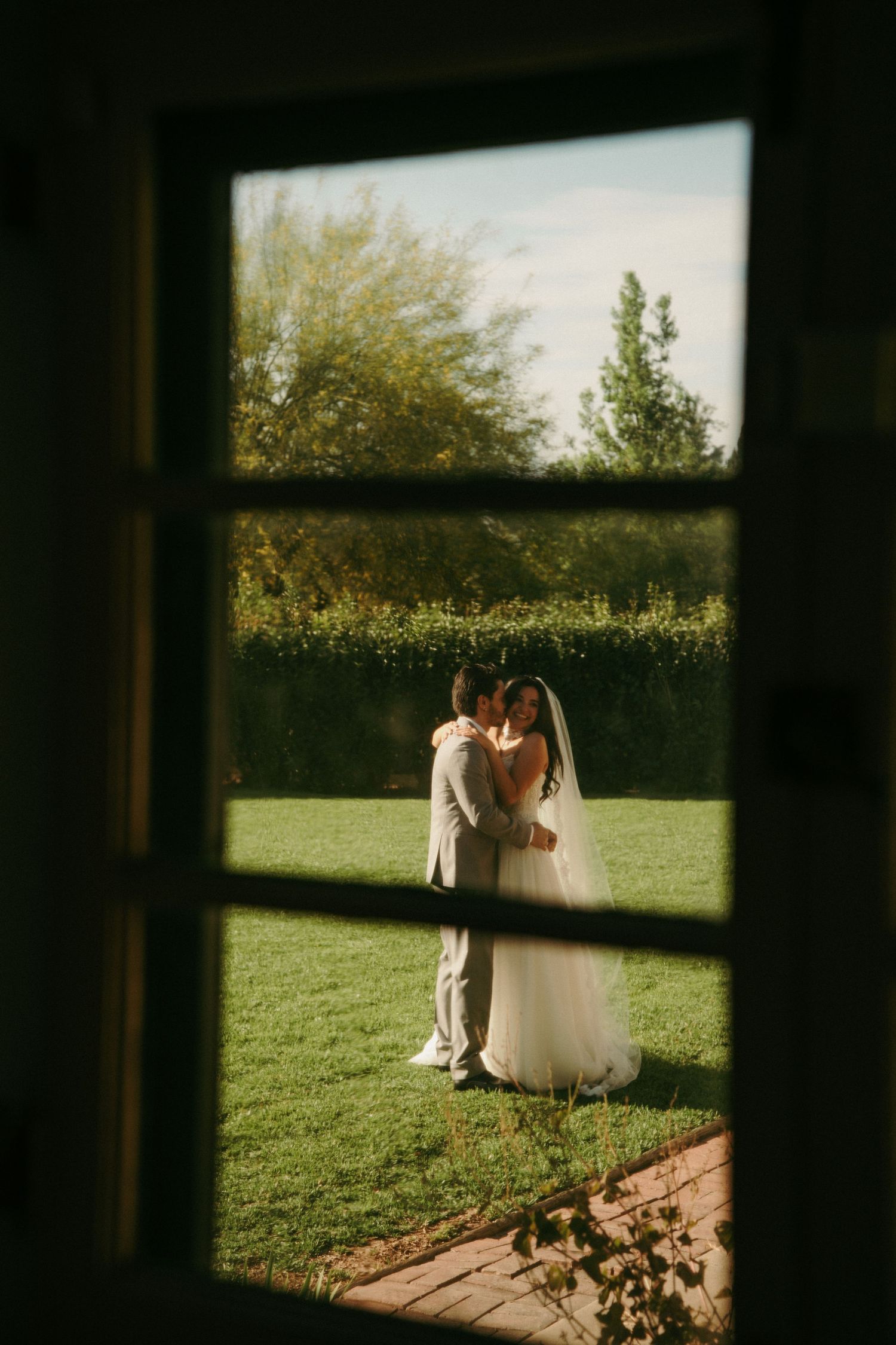 Bride and groom share a romantic kiss in a sunlit garden, viewed through a rustic window frame.