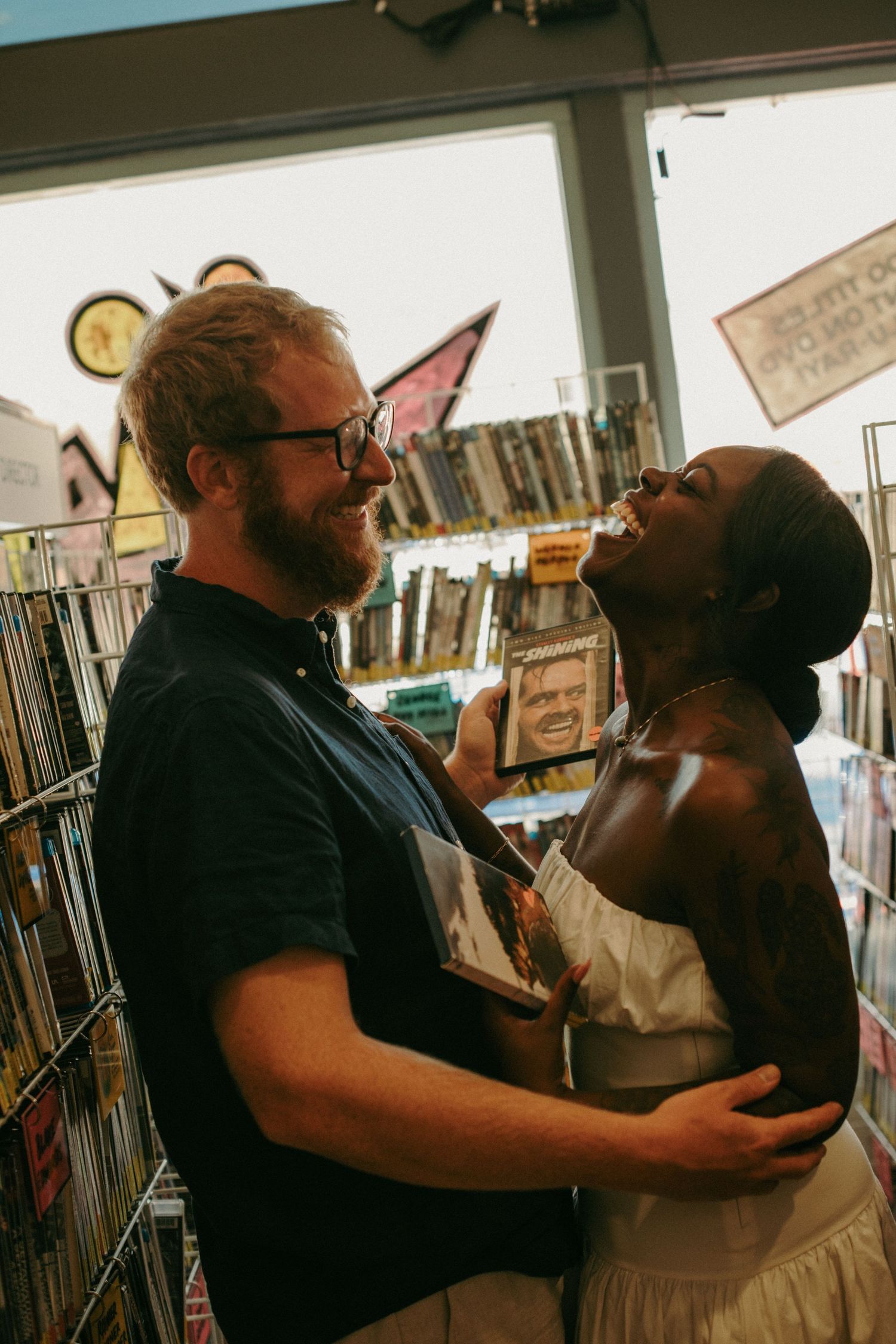 Couple laughing and embracing in a record store, holding vinyl albums among shelves of music.