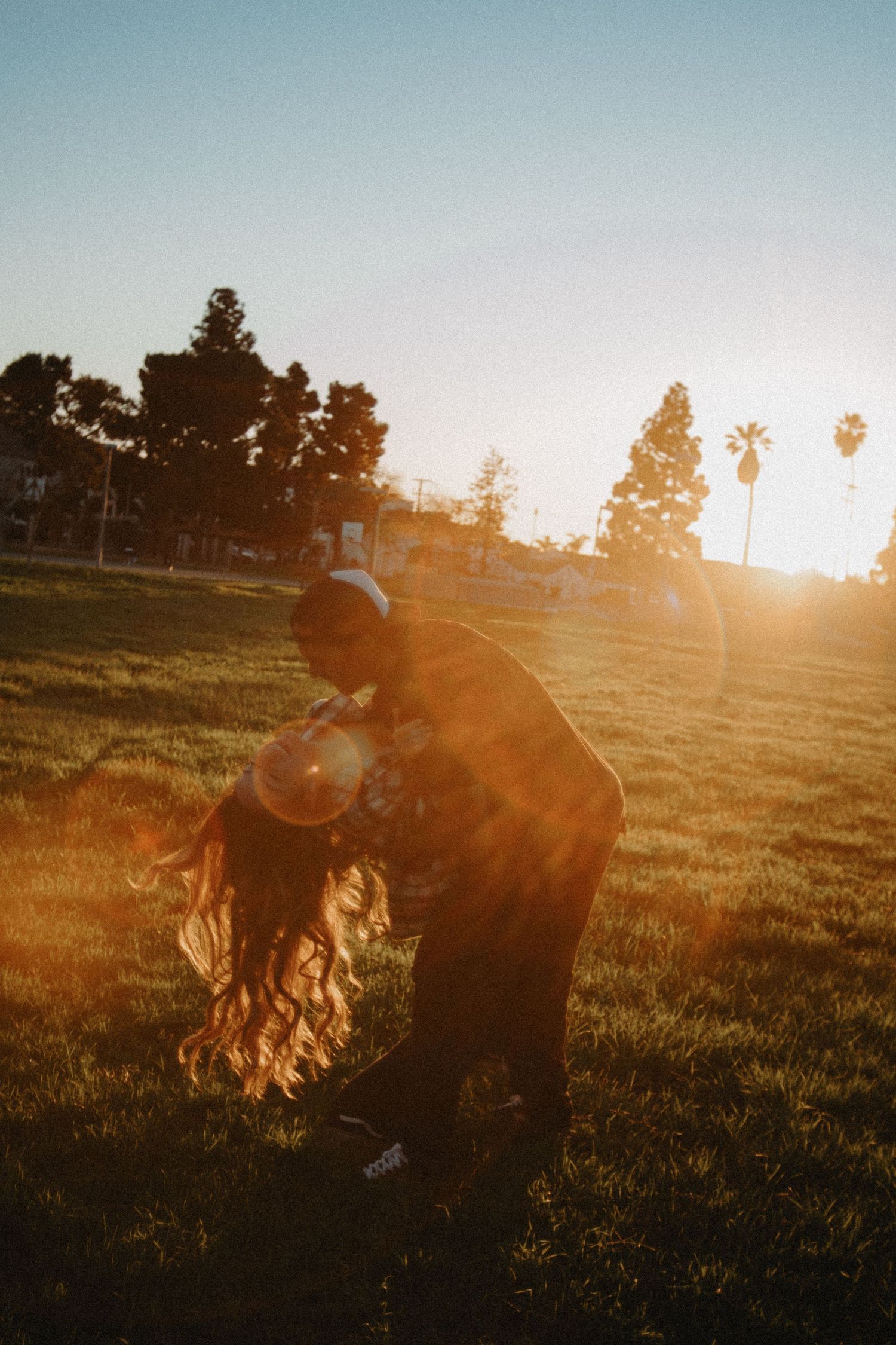 Couple dancing in golden sunset light on green grass field with palm trees in background.