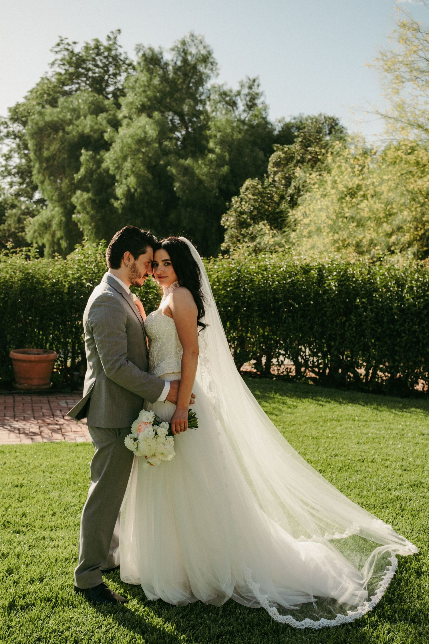 Bride and groom embrace on green lawn surrounded by lush hedges and trees on their wedding day.