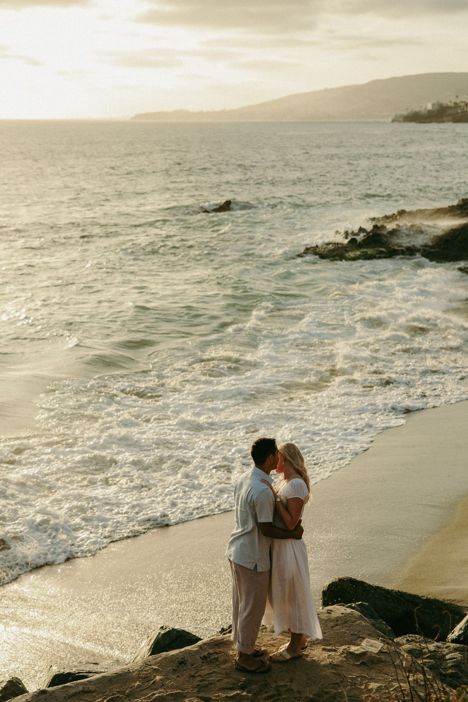 Couple sharing a romantic kiss on rocky shoreline at golden sunset with ocean waves and mountain backdrop.