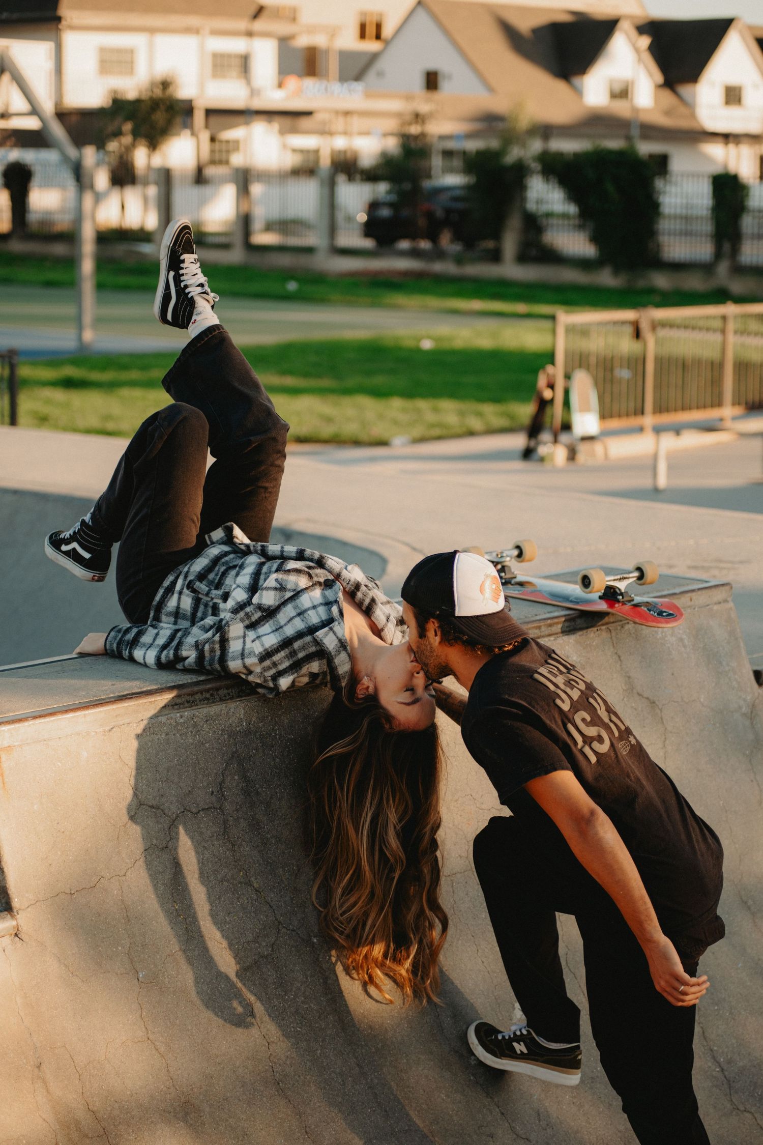 A couple shares a romantic kiss at a skate park, with the girl lying on a ramp and a skateboard resting nearby.