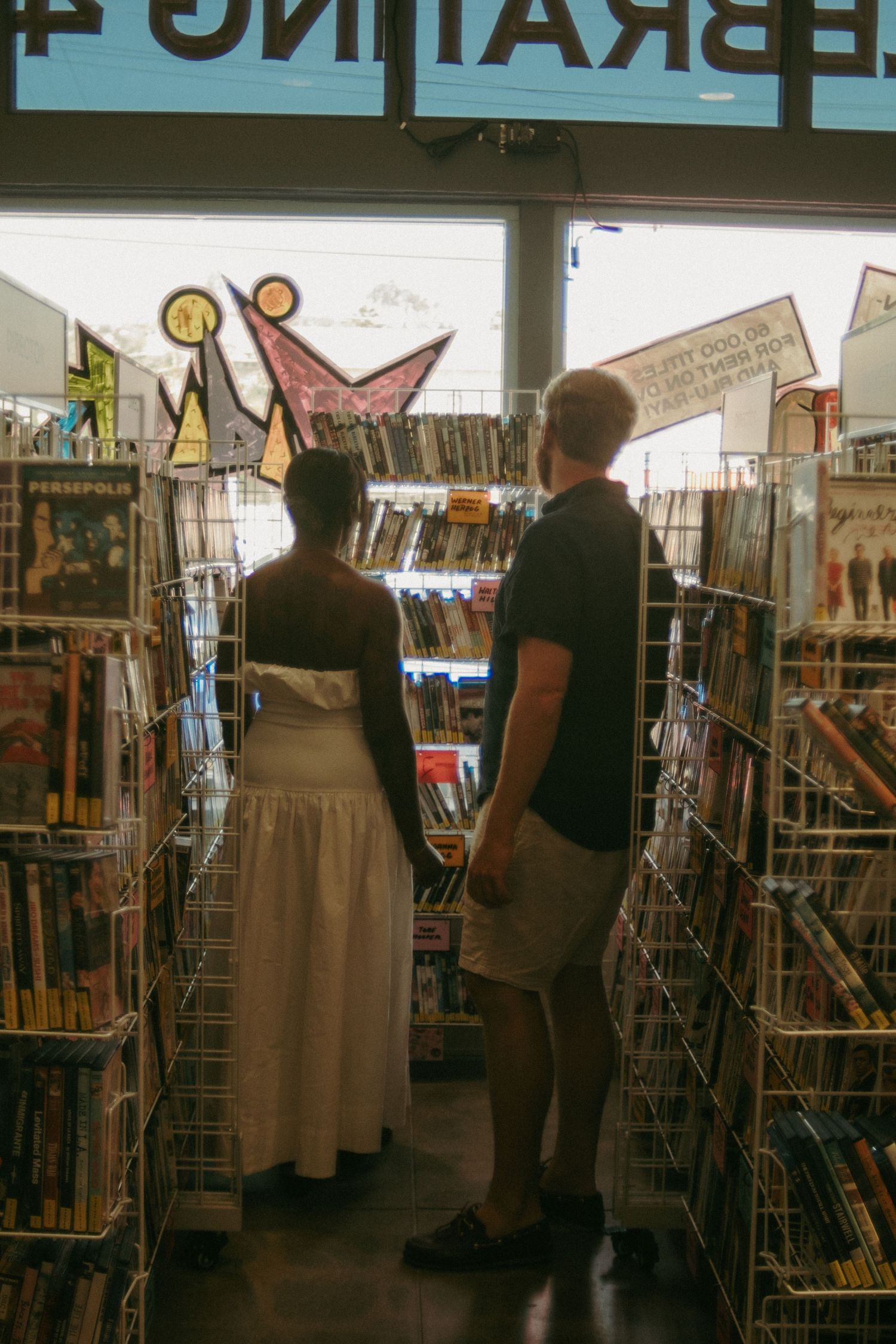 Couple browsing comic books and graphic novels in a bookstore aisle, woman in white dress, man in black shirt.