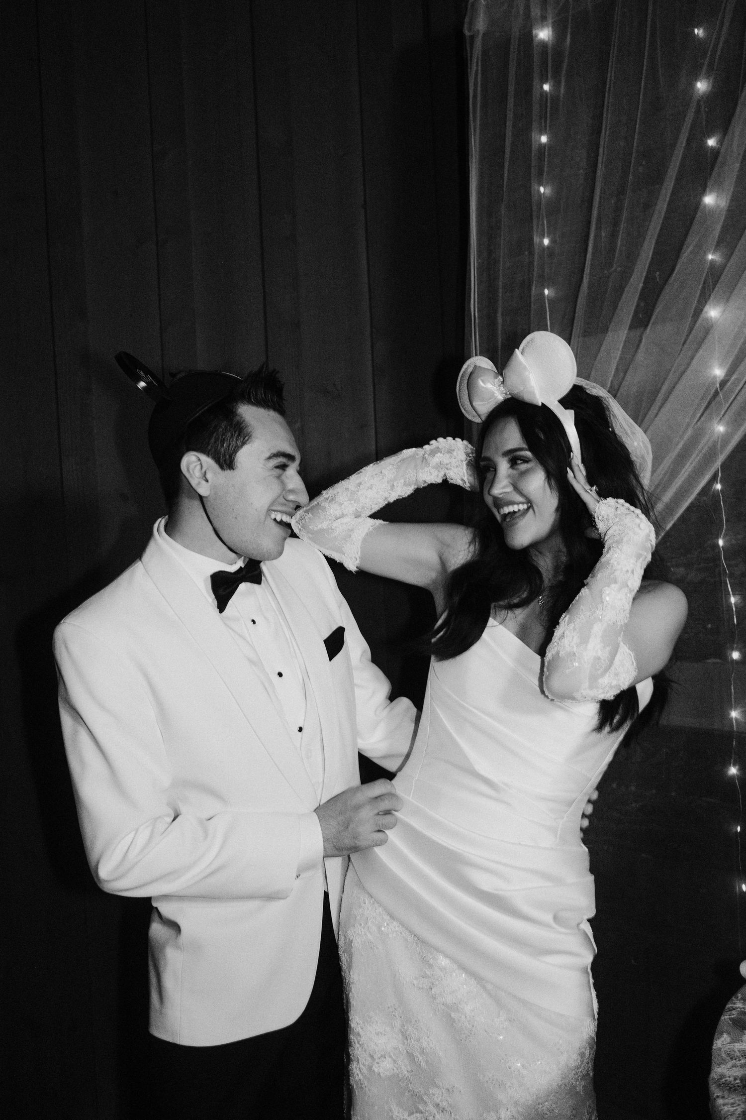 Black and white photo of a laughing bride and groom in formal wear with novelty ears posing before fairy lights.