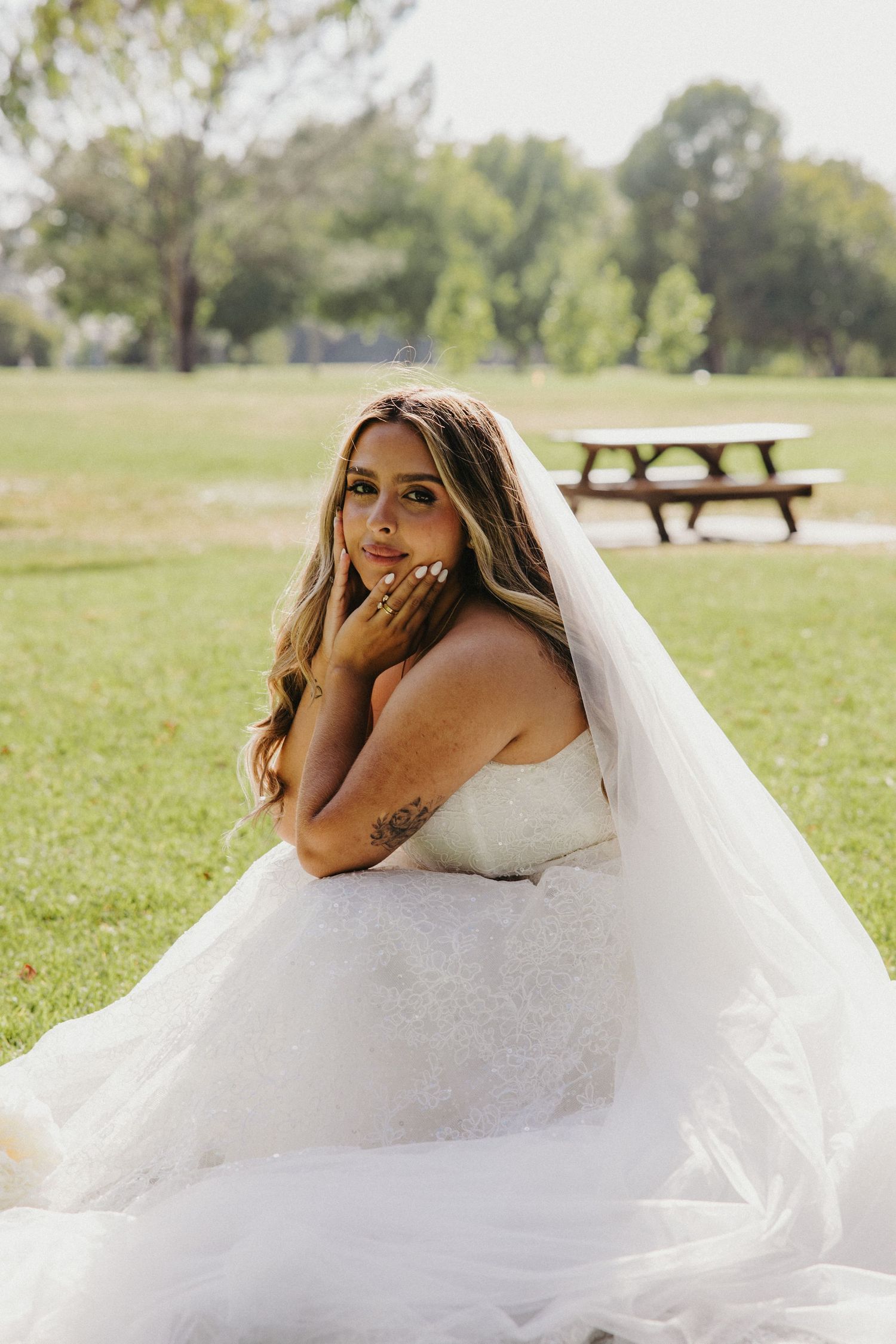 Bride in white gown and veil sits gracefully on green grass in a park, resting her face on her hand.