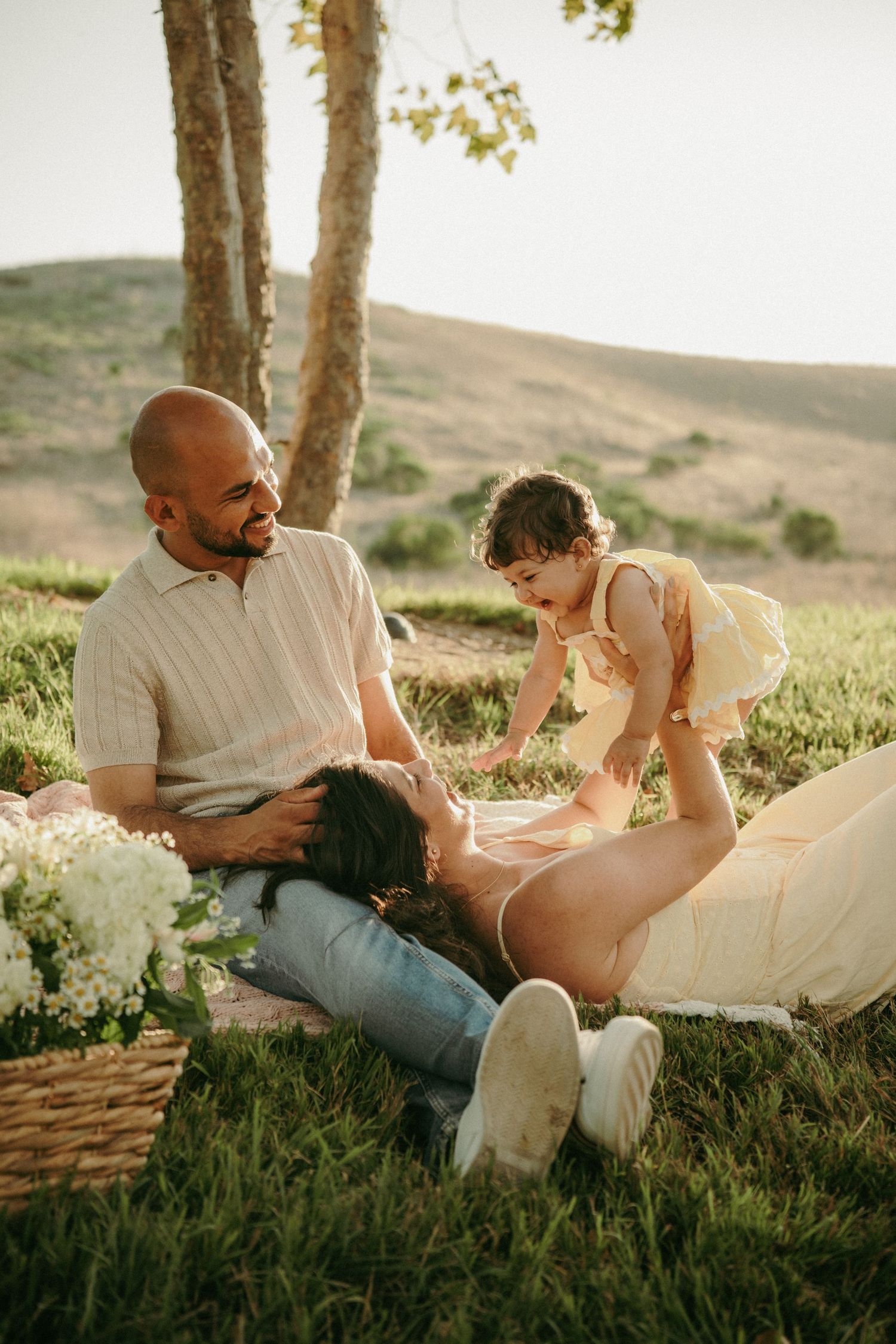 Family enjoying a sunny outdoor picnic on grass, with a bald father, reclining mother, and playful toddler near trees.