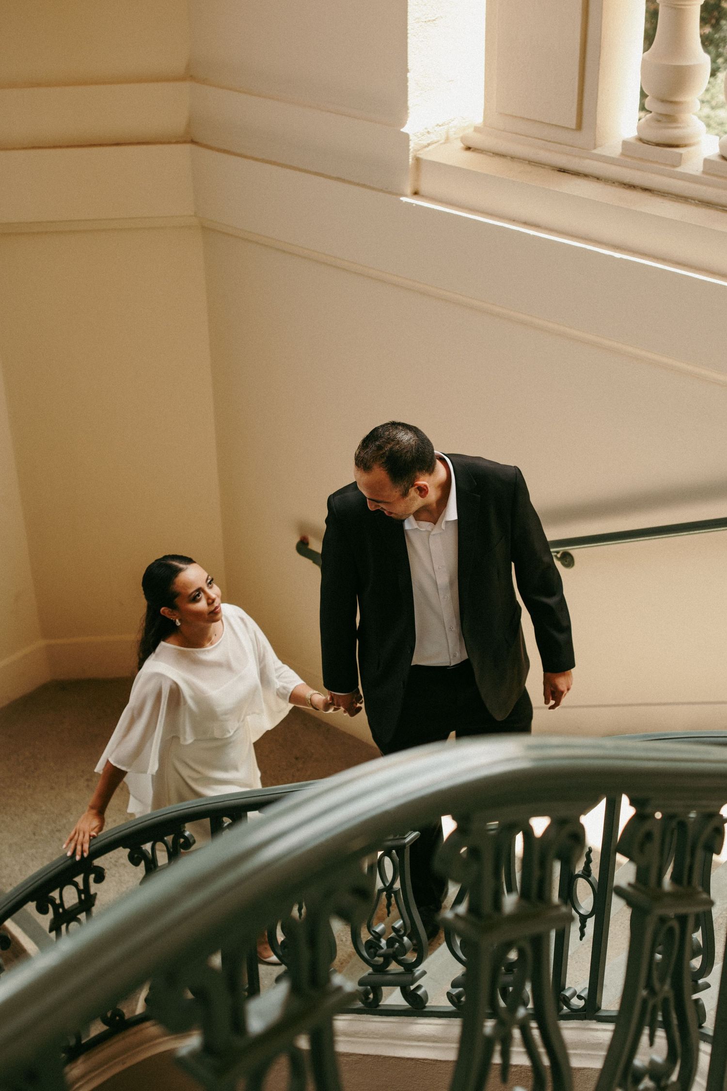 Elegant couple holding hands walking up ornate staircase in classic building.
