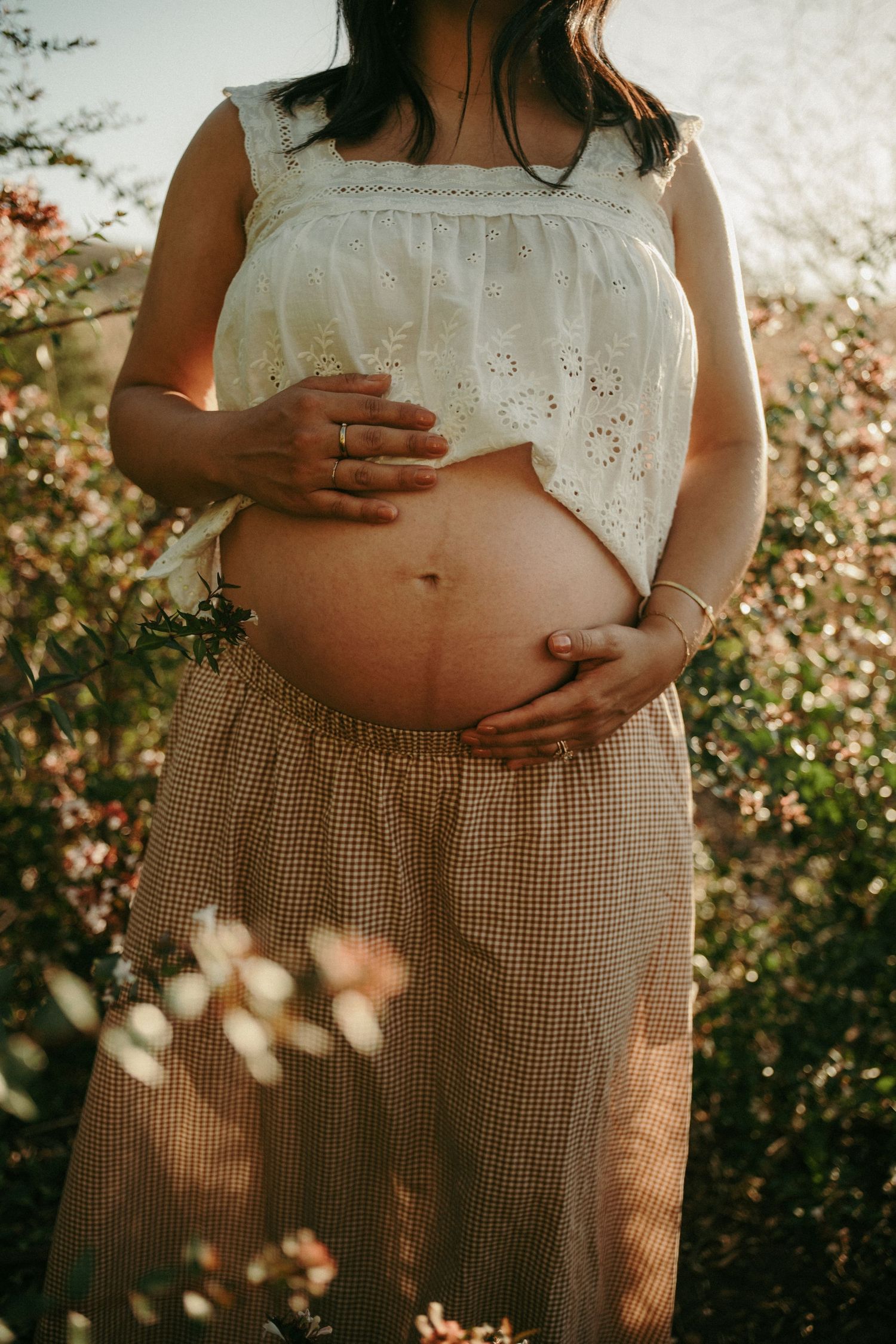 Pregnant woman outdoors holding baby bump, wearing white eyelet top and gingham skirt among flowers.