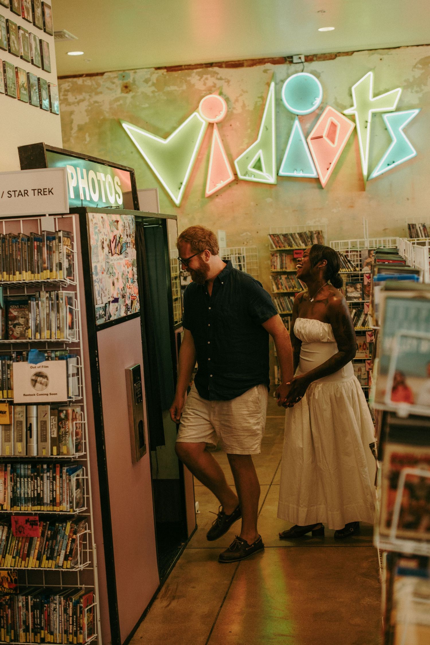 Couple holding hands in Vidiots video store, woman in white dress and man in blue shirt browsing shelves.