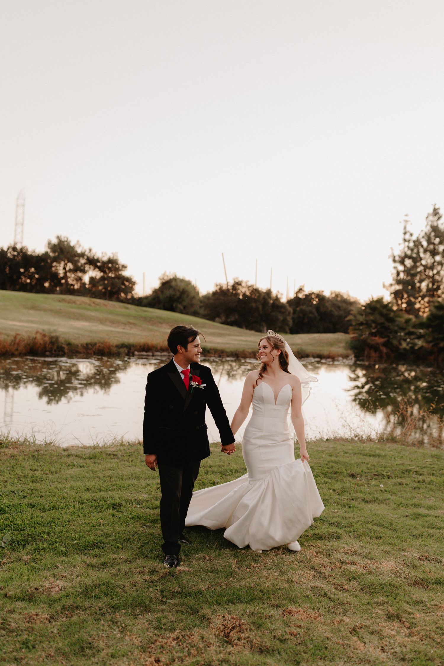 Bride and groom hold hands walking by a pond at golden hour, bride in mermaid gown with veil, groom in black tuxedo.
