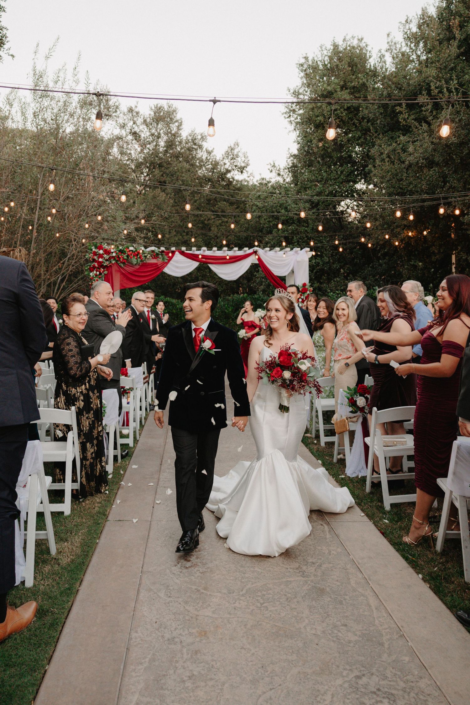 Newlywed couple walks down outdoor wedding aisle amid cheering guests, string lights, and red floral decor.