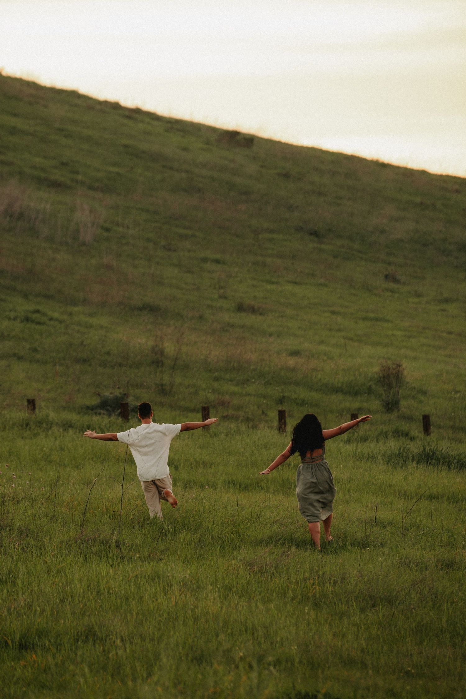 Two people running freely through a lush green hillside meadow at sunset with arms outstretched.