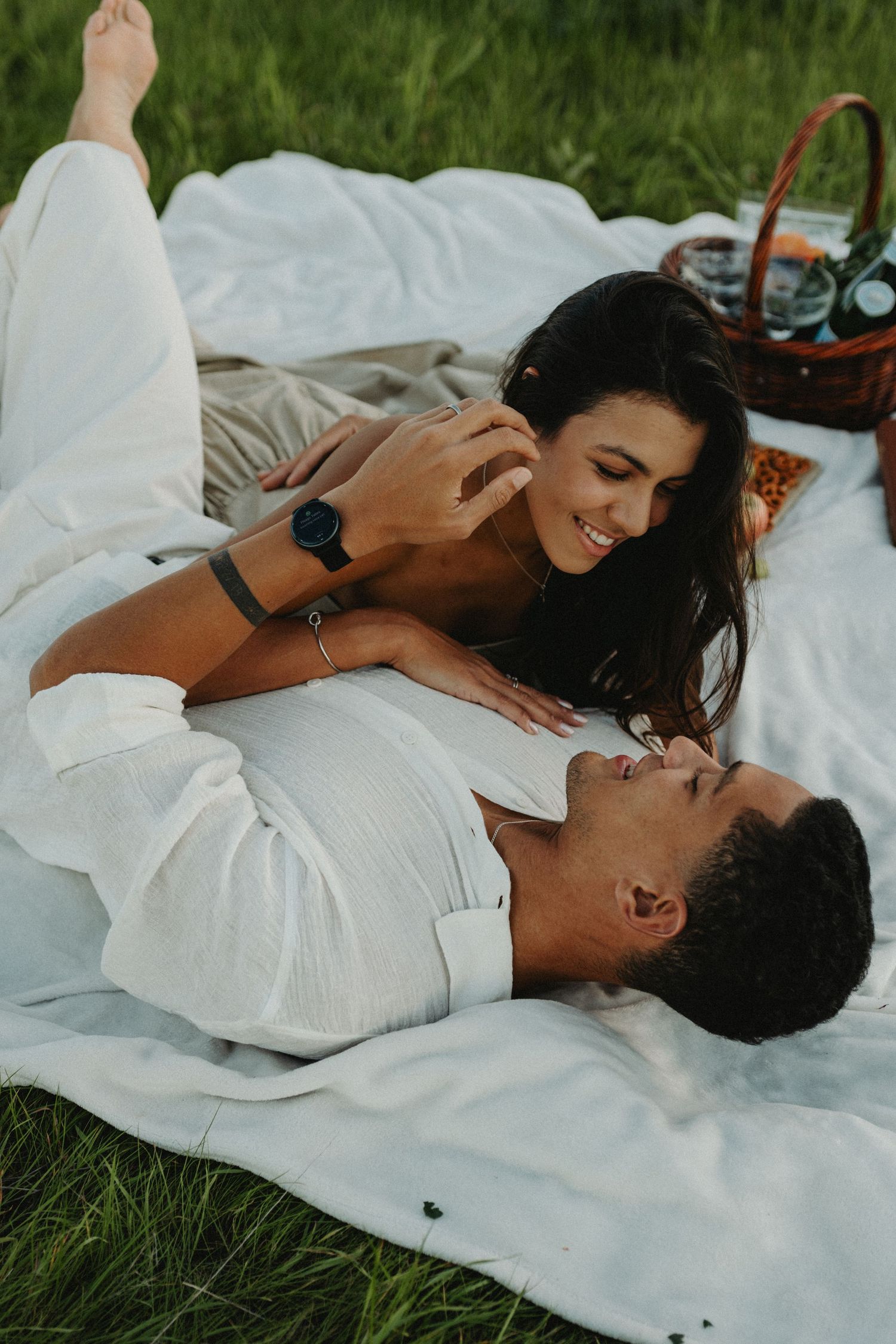 Couple sharing a romantic picnic on white blanket in green field, smiling and gazing at each other.