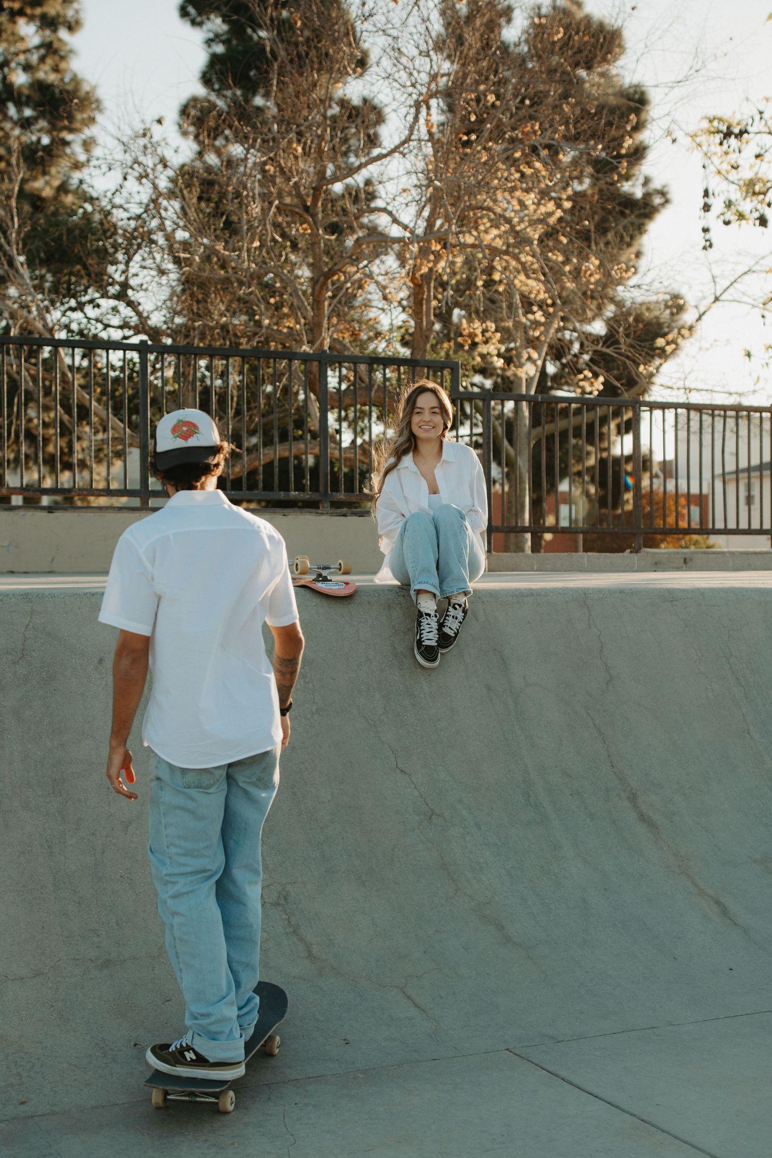 A young couple at a skate park, man on skateboard facing woman sitting on concrete ramp, surrounded by trees.