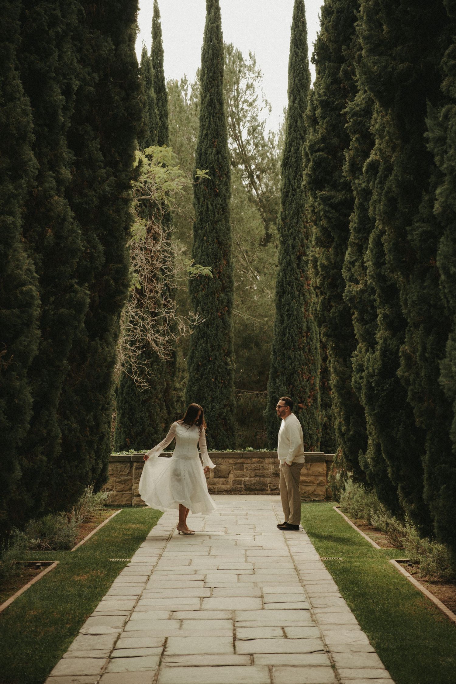 Couple in wedding attire walking along a stone pathway lined with tall cypress trees in a lush garden.
