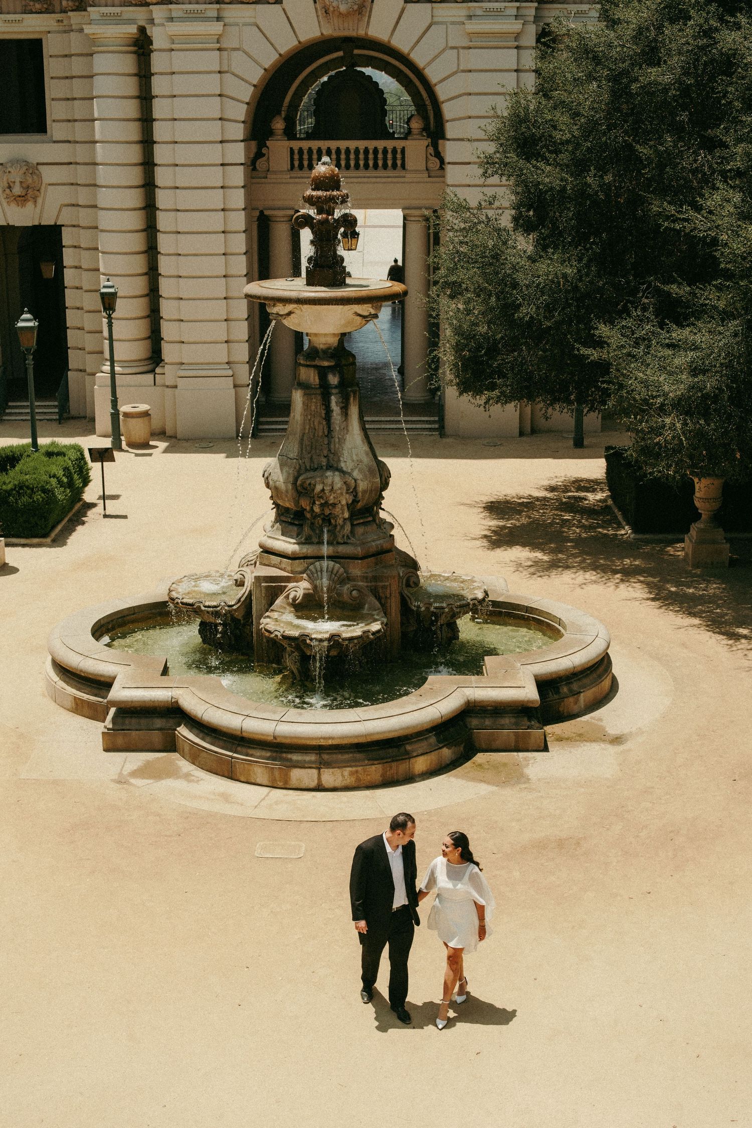 Couple walks hand in hand before an ornate stone fountain in a sunlit courtyard of a grand historic building.