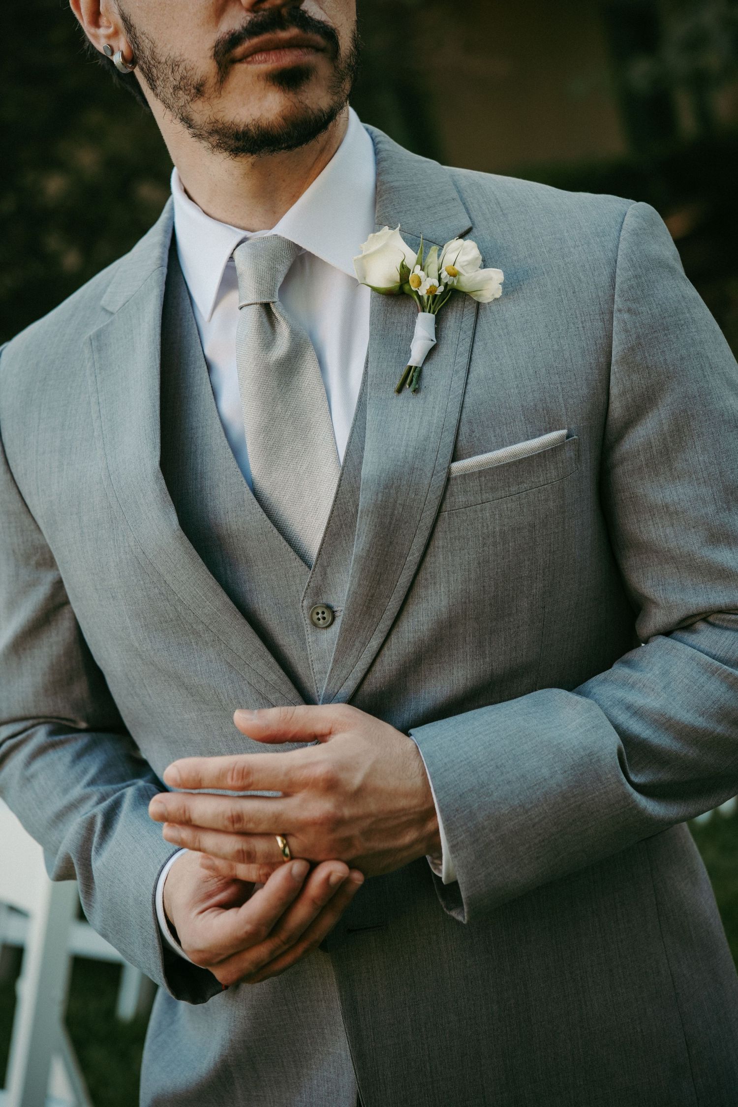 Groom in gray three-piece suit adjusting cufflinks, wearing gold wedding band and white floral boutonniere.
