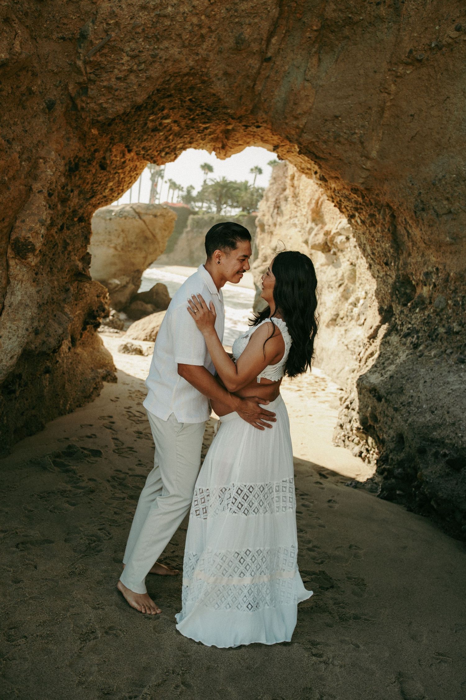 Couple in white embracing under a rocky beach cave arch with palm trees in the background.