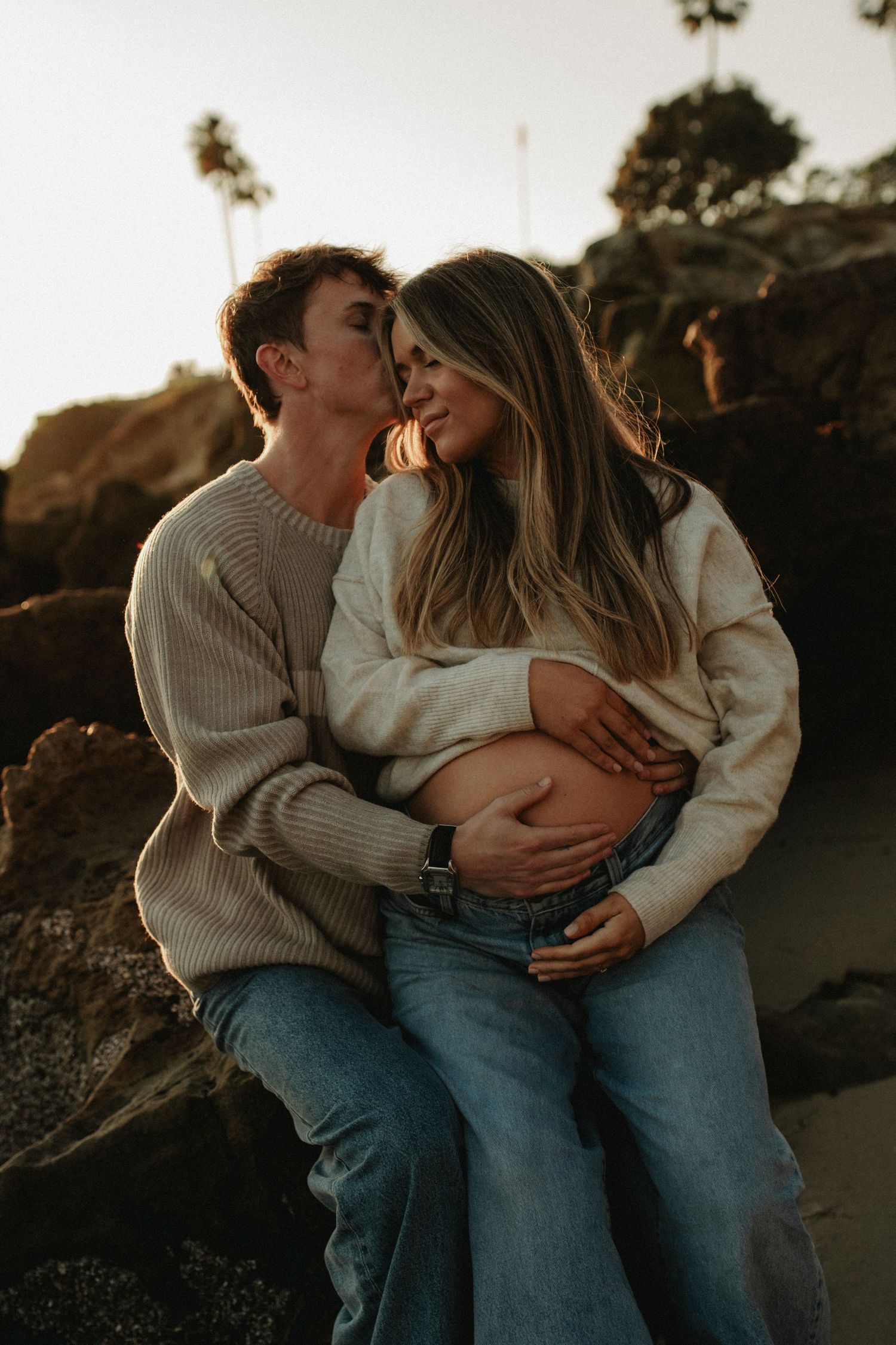 Romantic couple sharing a tender moment on rocky beach at sunset, cradling a baby bump.