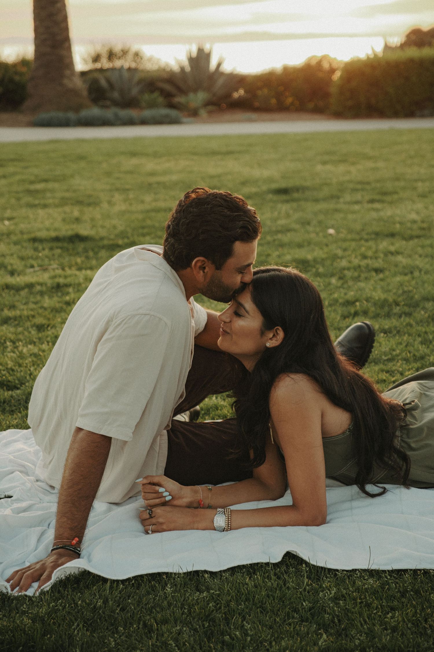 Couple sharing a tender kiss on a blanket in a sunlit park at golden hour.