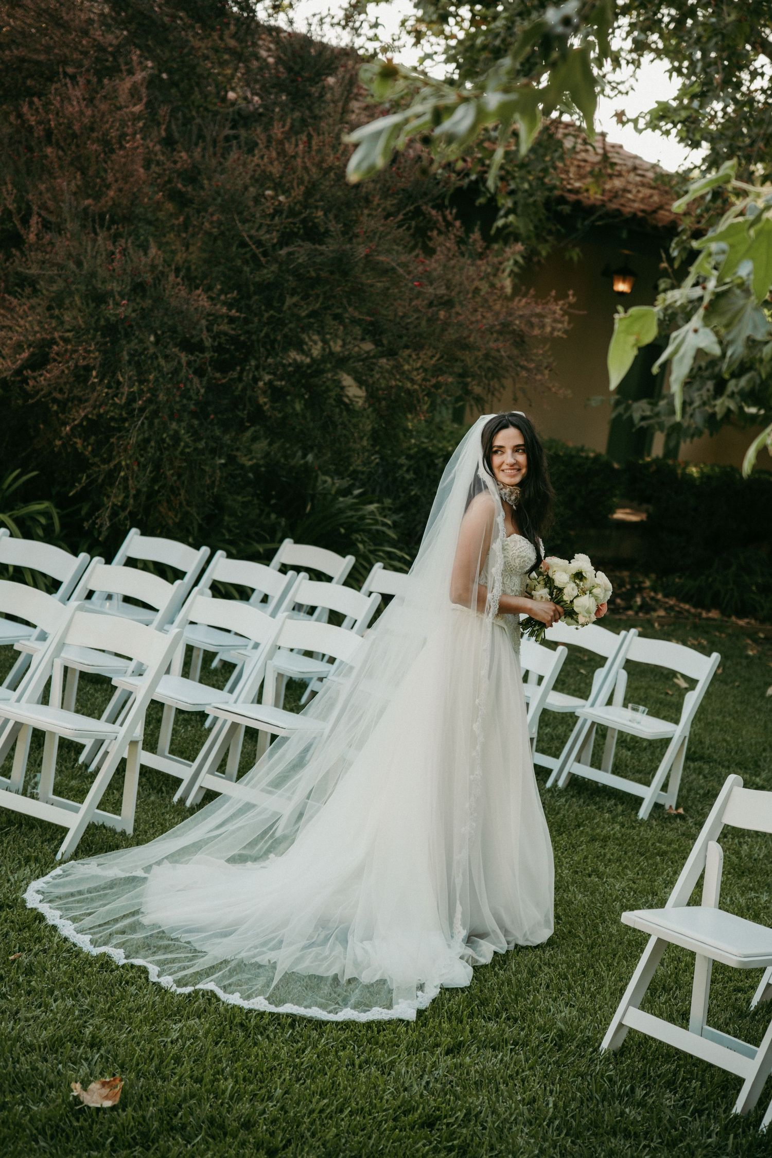 Smiling bride in white gown and veil holds bouquet outdoors among white chairs at garden wedding ceremony.