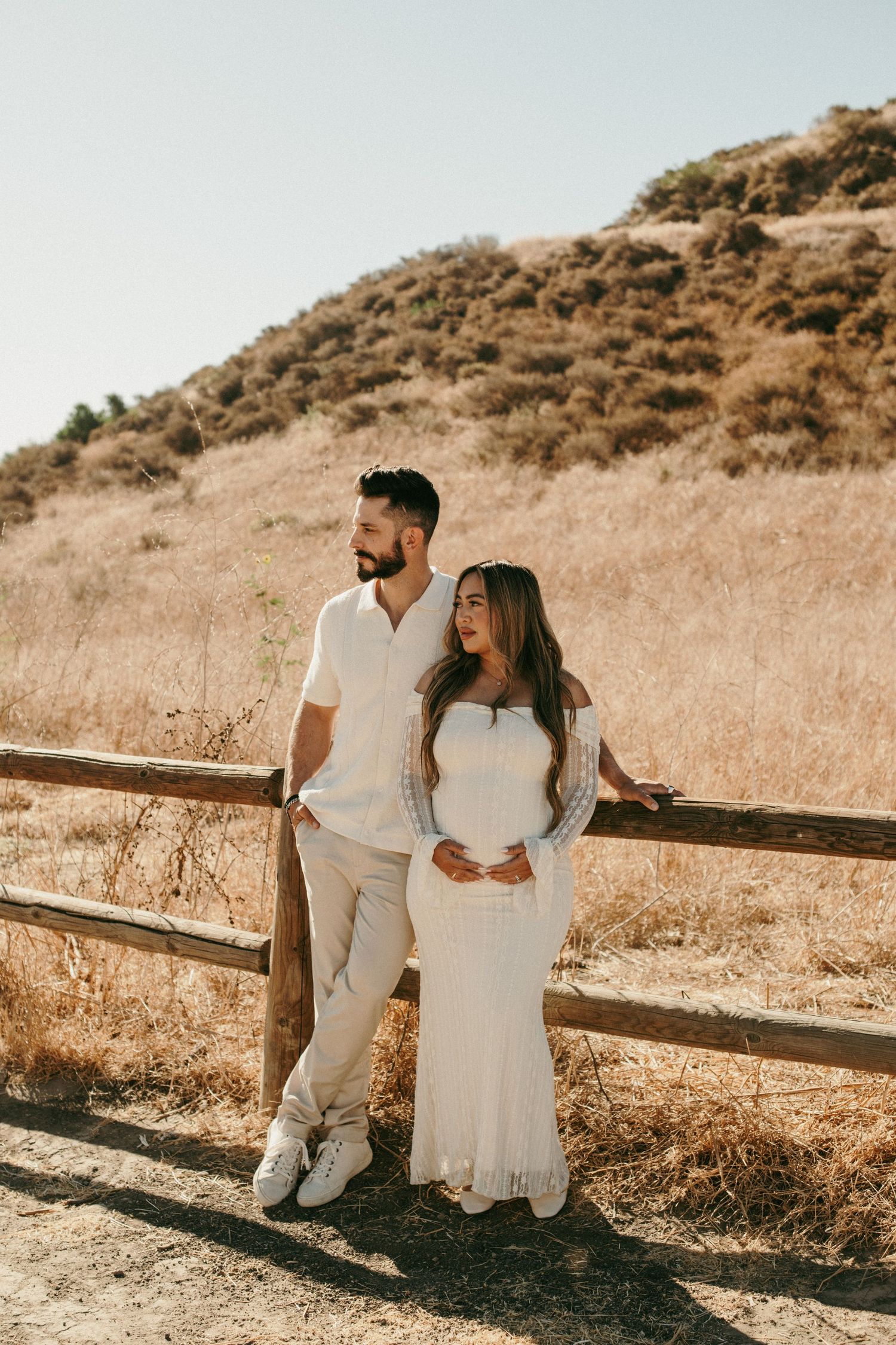 Couple in white outfits posing by a wooden fence with dry golden hills in the background on a sunny day.