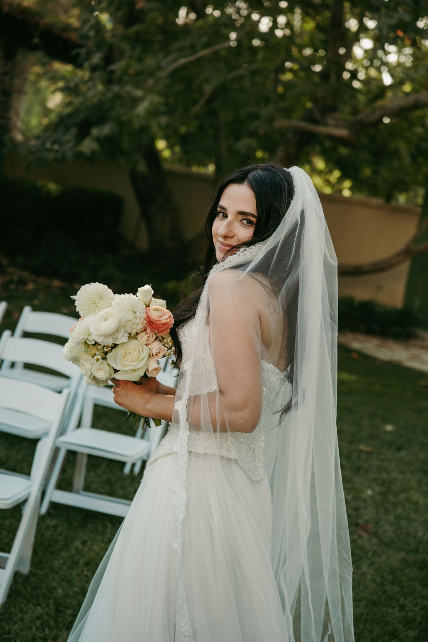 Bride in white gown and veil holds floral bouquet outdoors near white chairs, smiling over her shoulder.