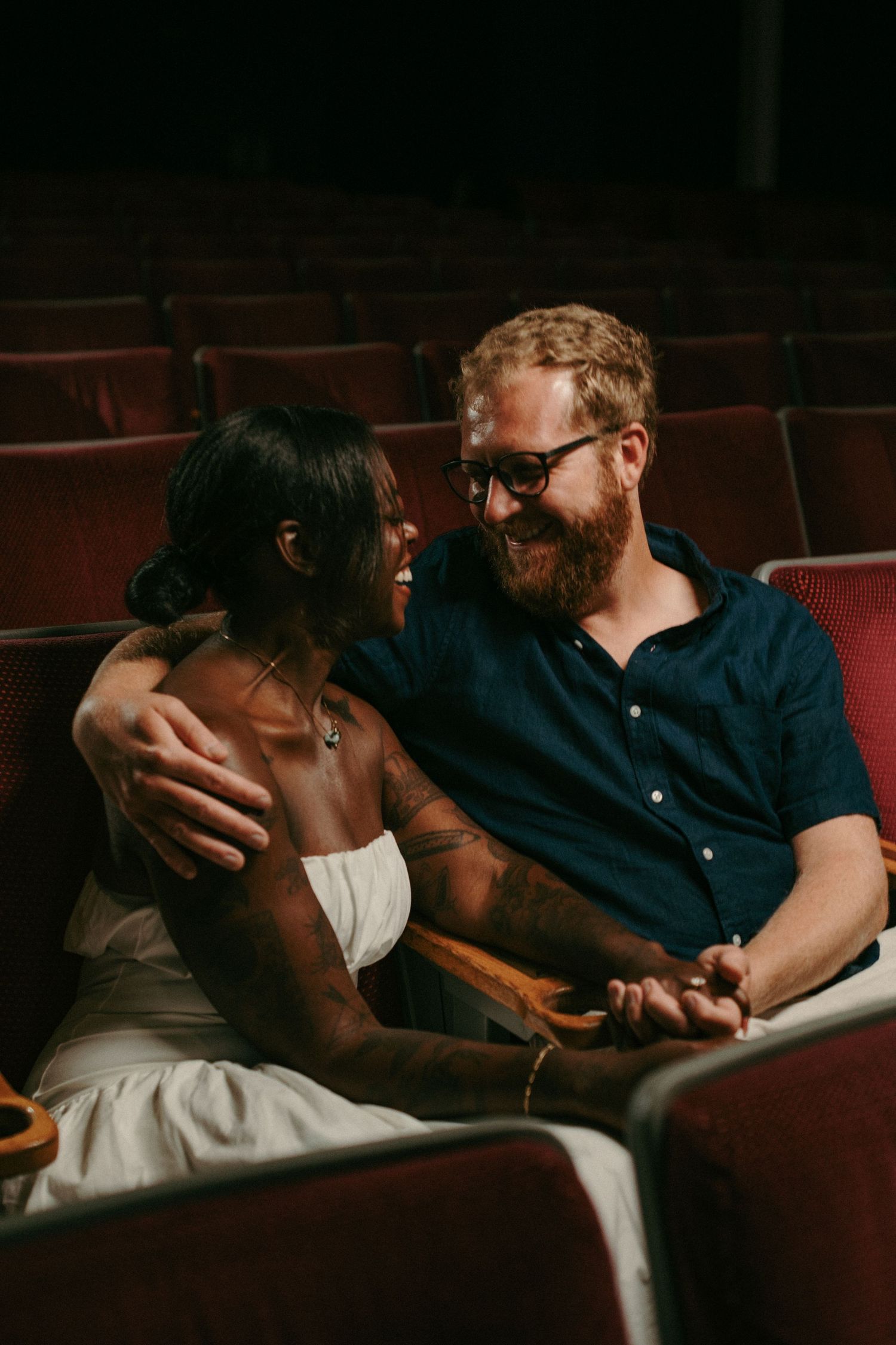 A happy interracial couple sits close together in vintage red theater seats, smiling and holding hands.