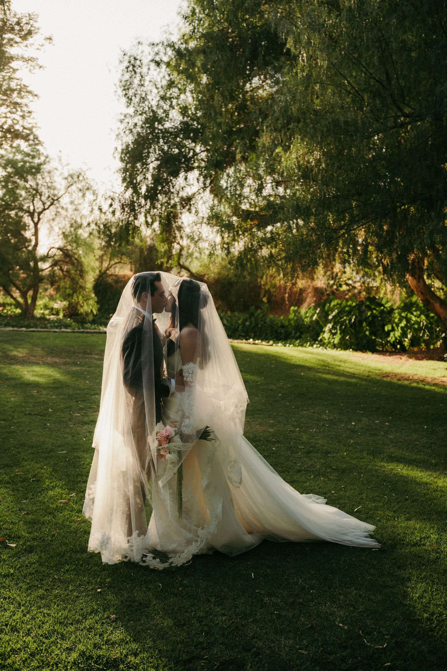 Two brides sharing a kiss under flowing veils on lush green lawn surrounded by trees on their wedding day.