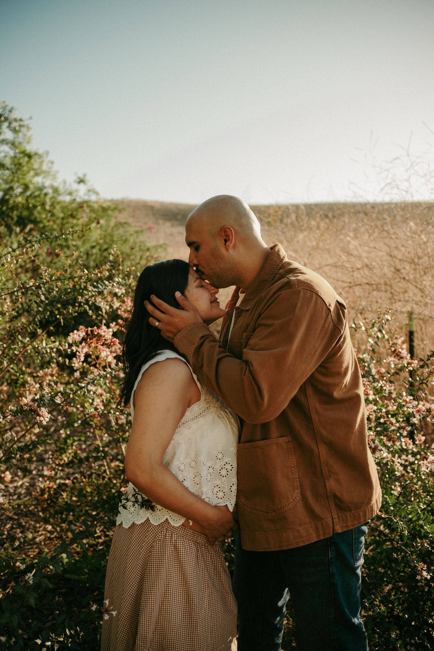 Couple sharing a tender kiss outdoors surrounded by blooming pink flowers in warm golden light.