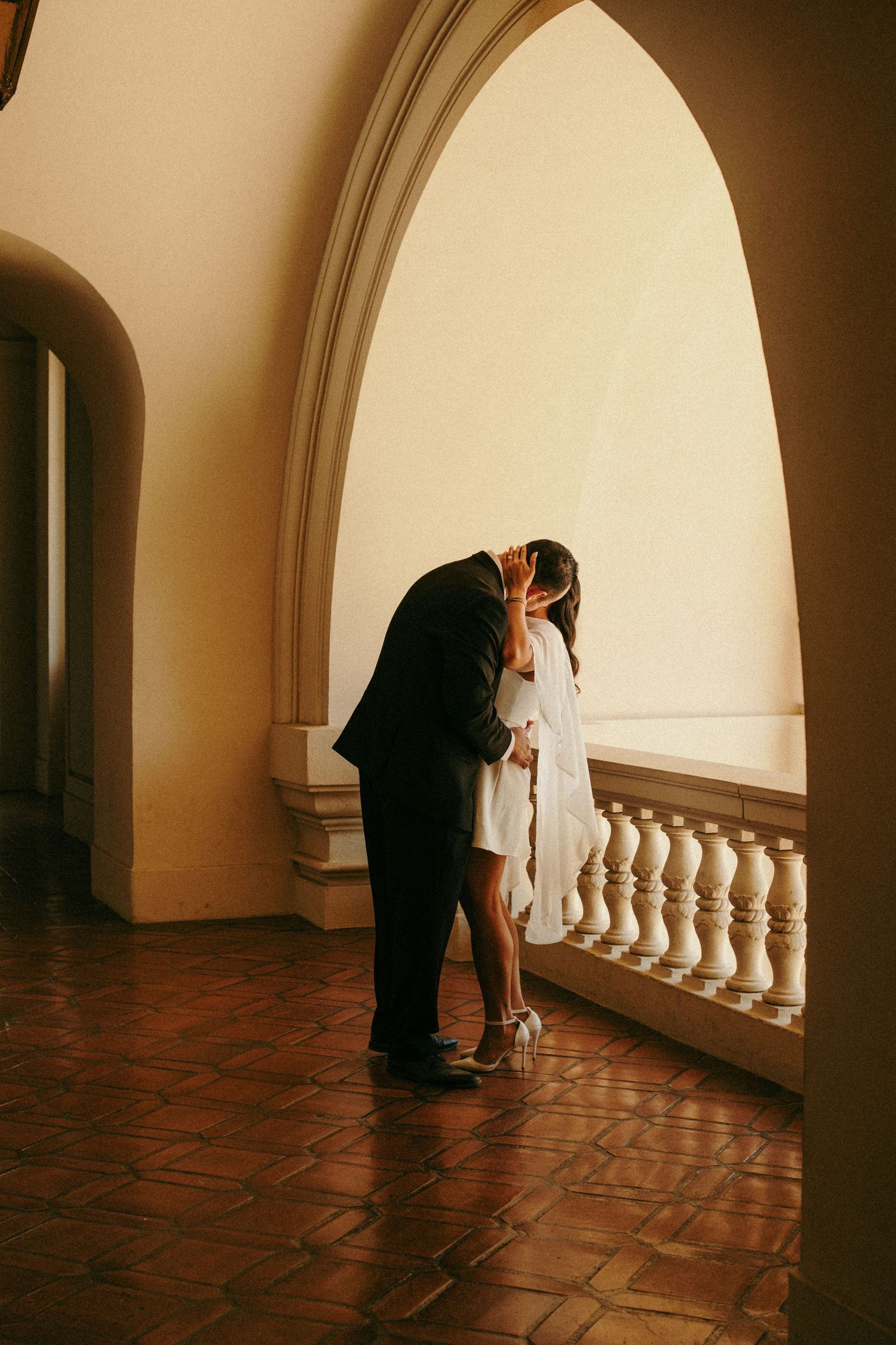 Couple sharing a romantic kiss on an elegant balcony with arched doorways and terracotta floors.