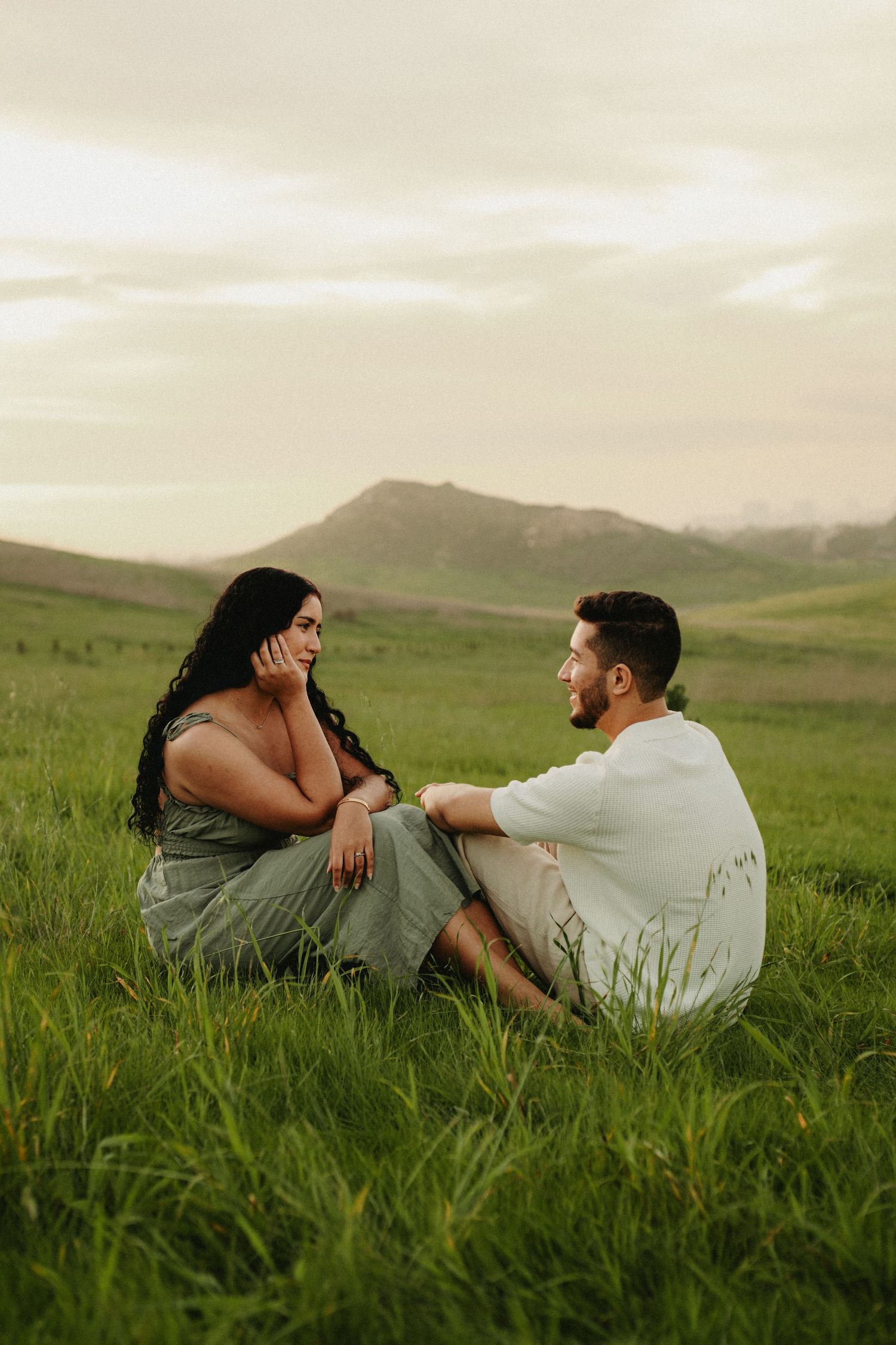 A couple sits together in a lush green meadow with rolling hills at golden hour.