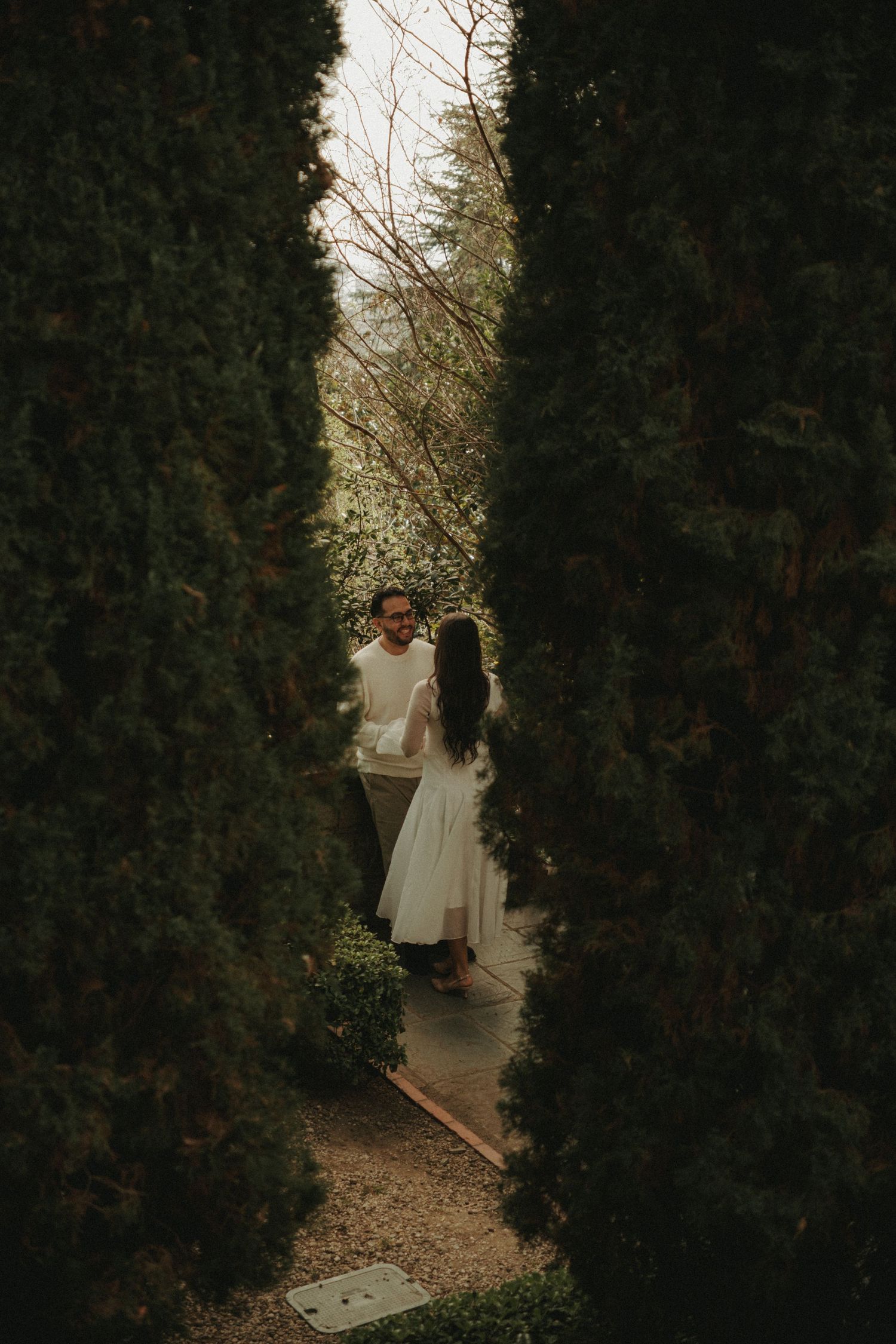 Couple in white outfits standing between tall cypress trees on a garden path, sharing a romantic moment.