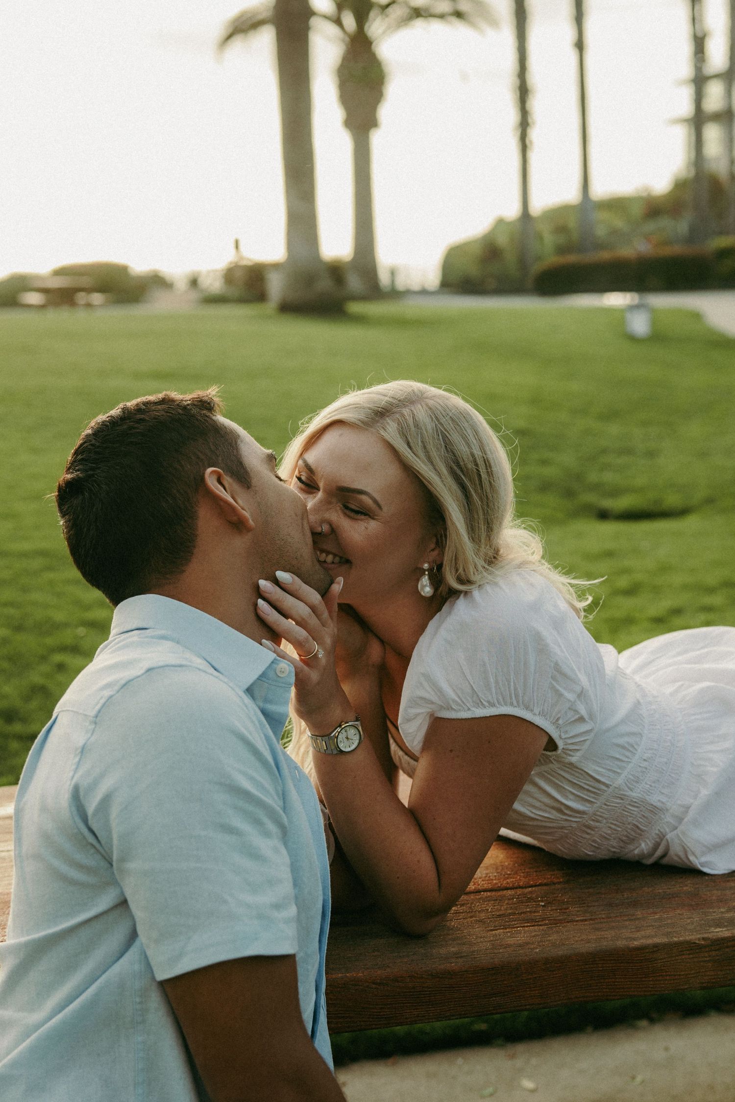 Blonde woman in white dress leaning in to kiss her partner while resting on a wooden bench outdoors.