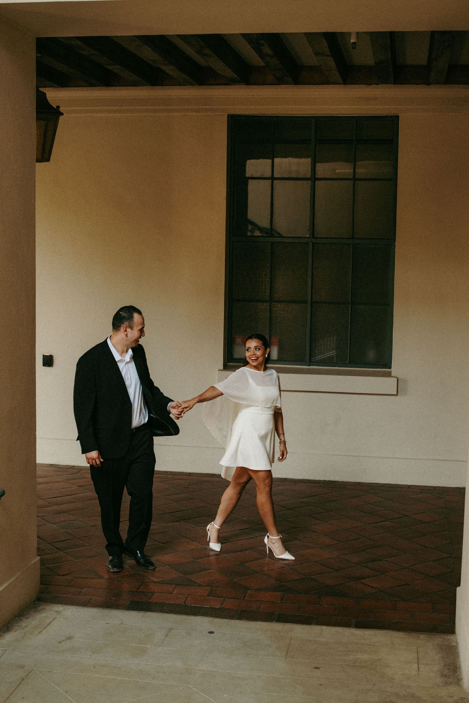 Couple in wedding attire holding hands and dancing playfully under a covered outdoor archway.
