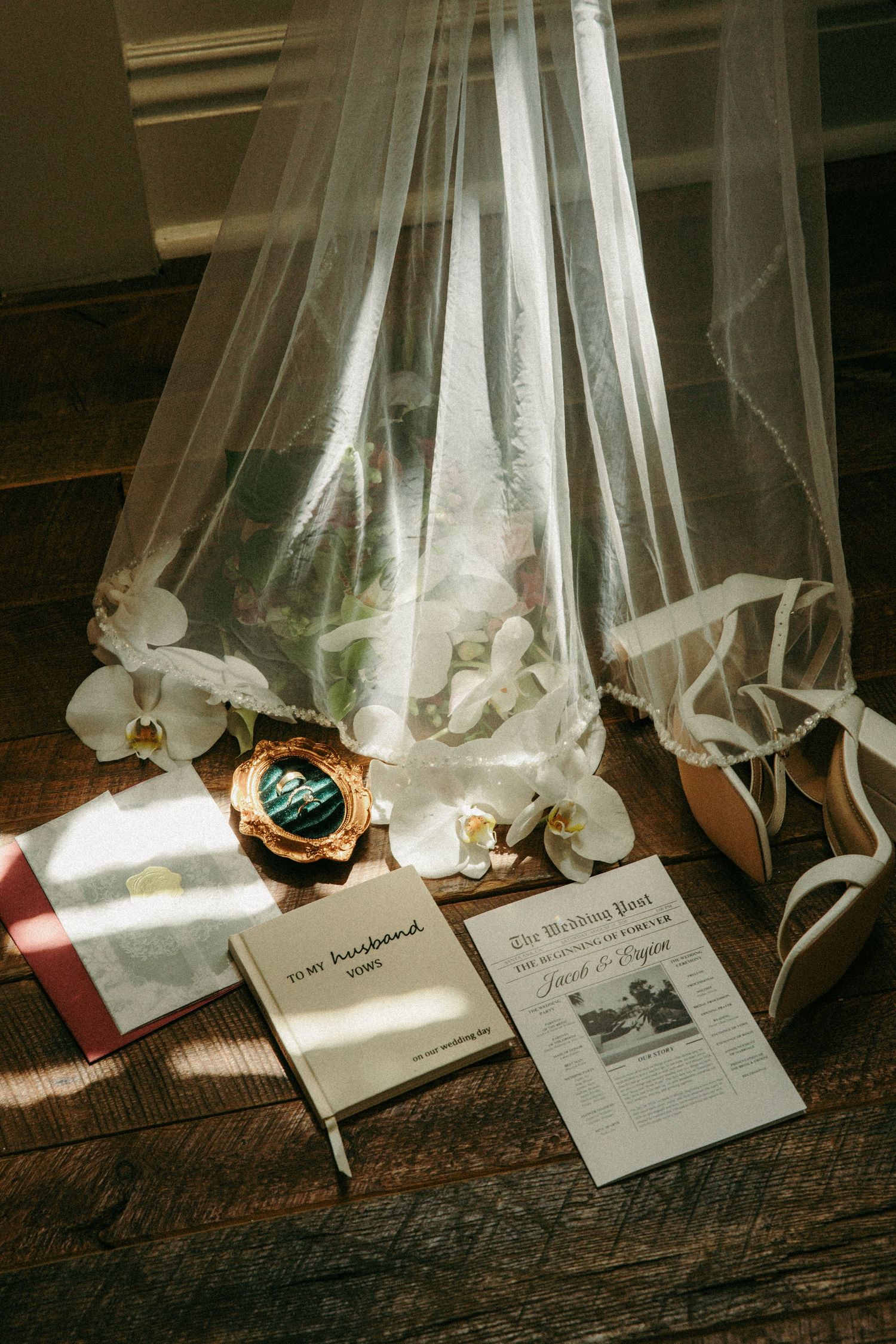 Wedding flat lay with veil, bridal bouquet, vow books, and wedding newspaper on wood floor.