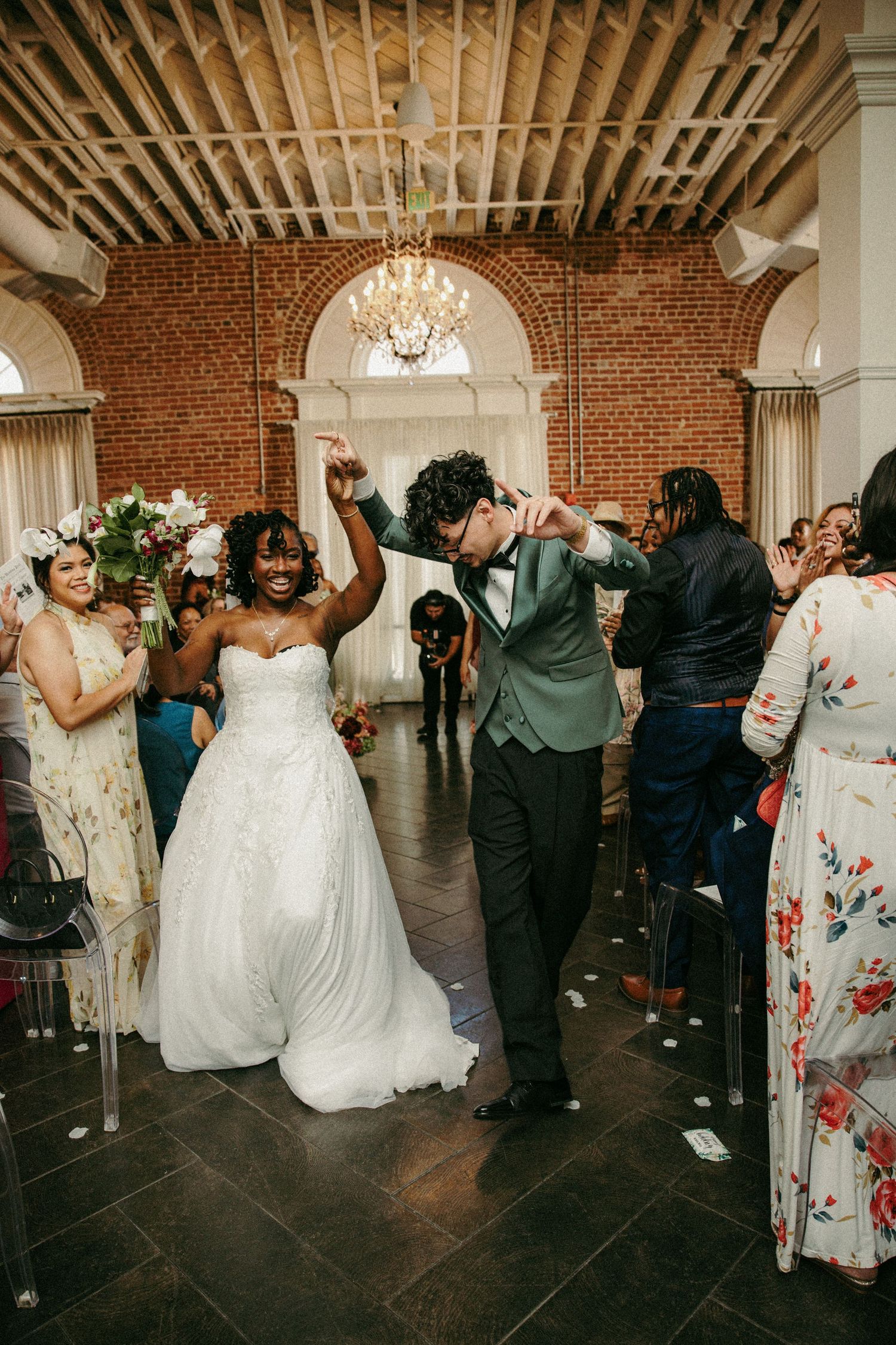 Joyful newlyweds dance down the aisle at their elegant brick-walled wedding venue, surrounded by cheering guests.