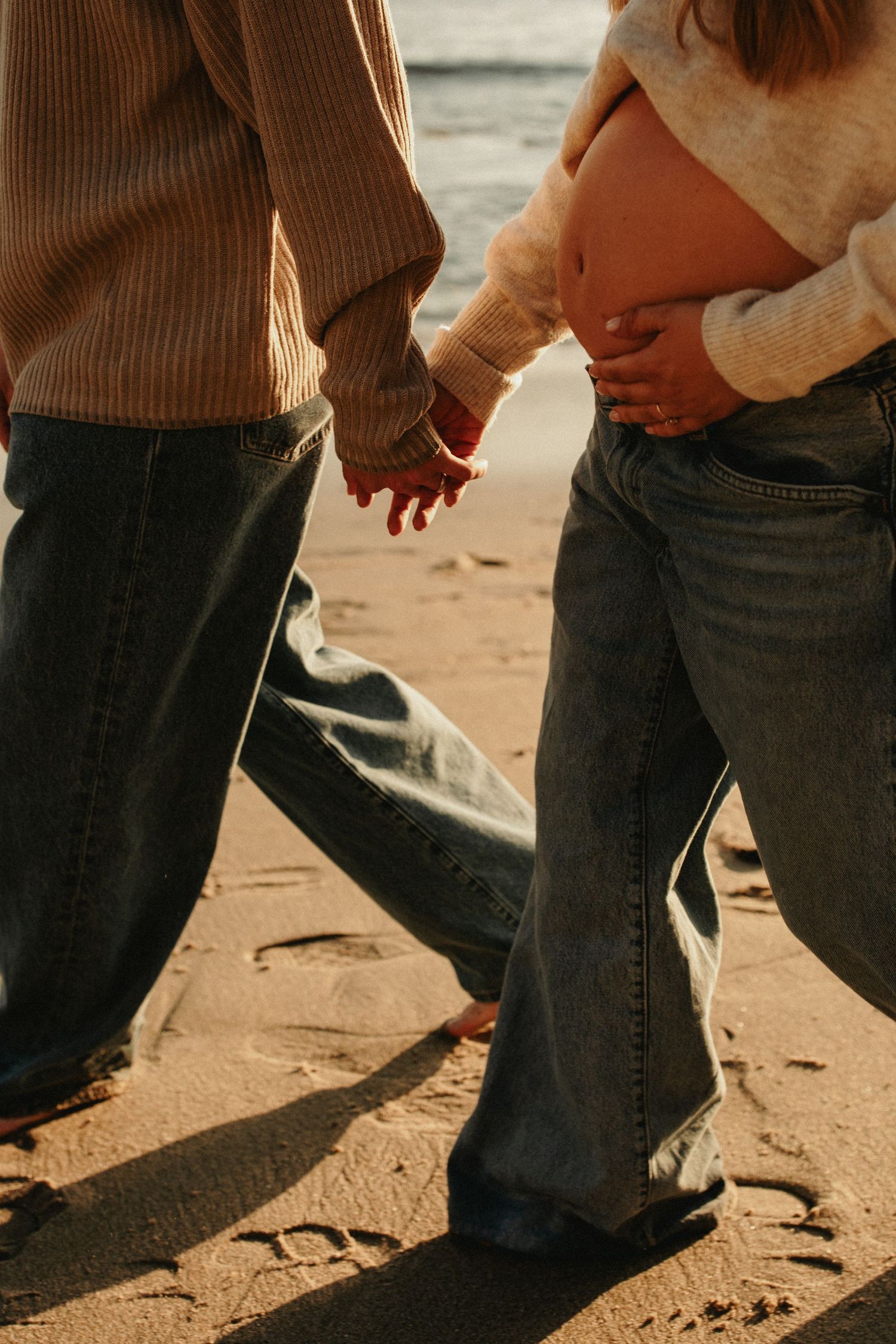 Pregnant couple holding hands on sandy beach, woman cradling baby bump, both wearing jeans and sweaters in golden sunlight.