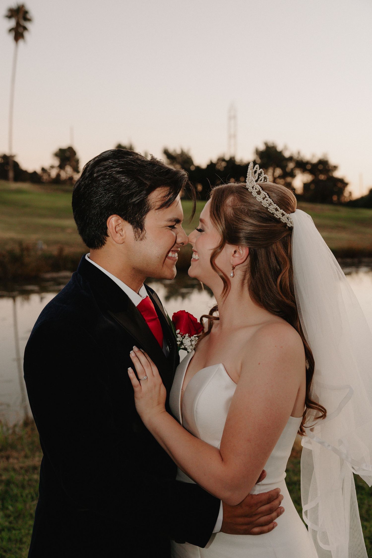 Bride and groom share a romantic moment, nearly kissing, with bride wearing a tiara and veil outdoors.