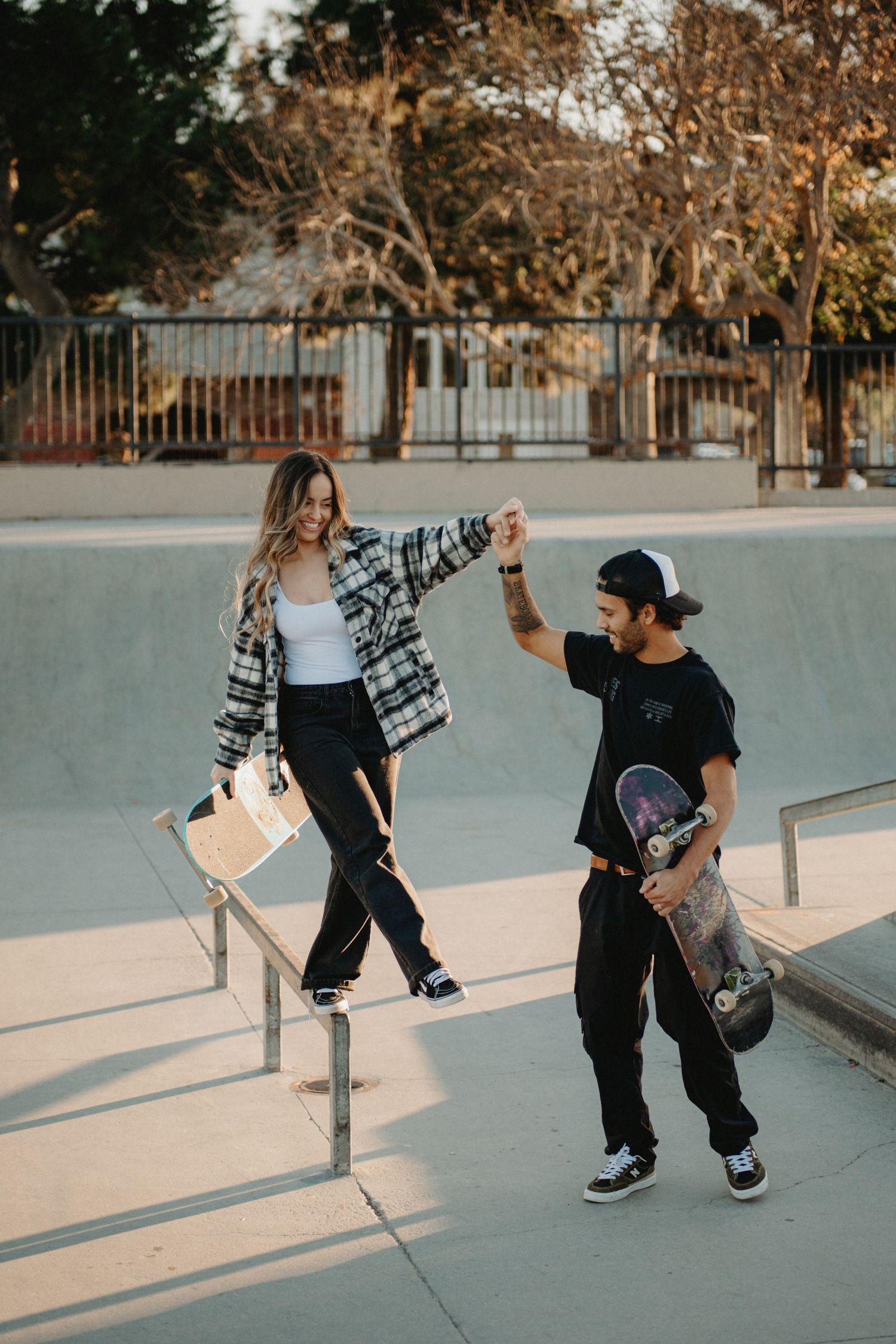 A couple holds hands at a skate park, woman balancing on a rail while man holds a skateboard, both wearing casual streetwear.