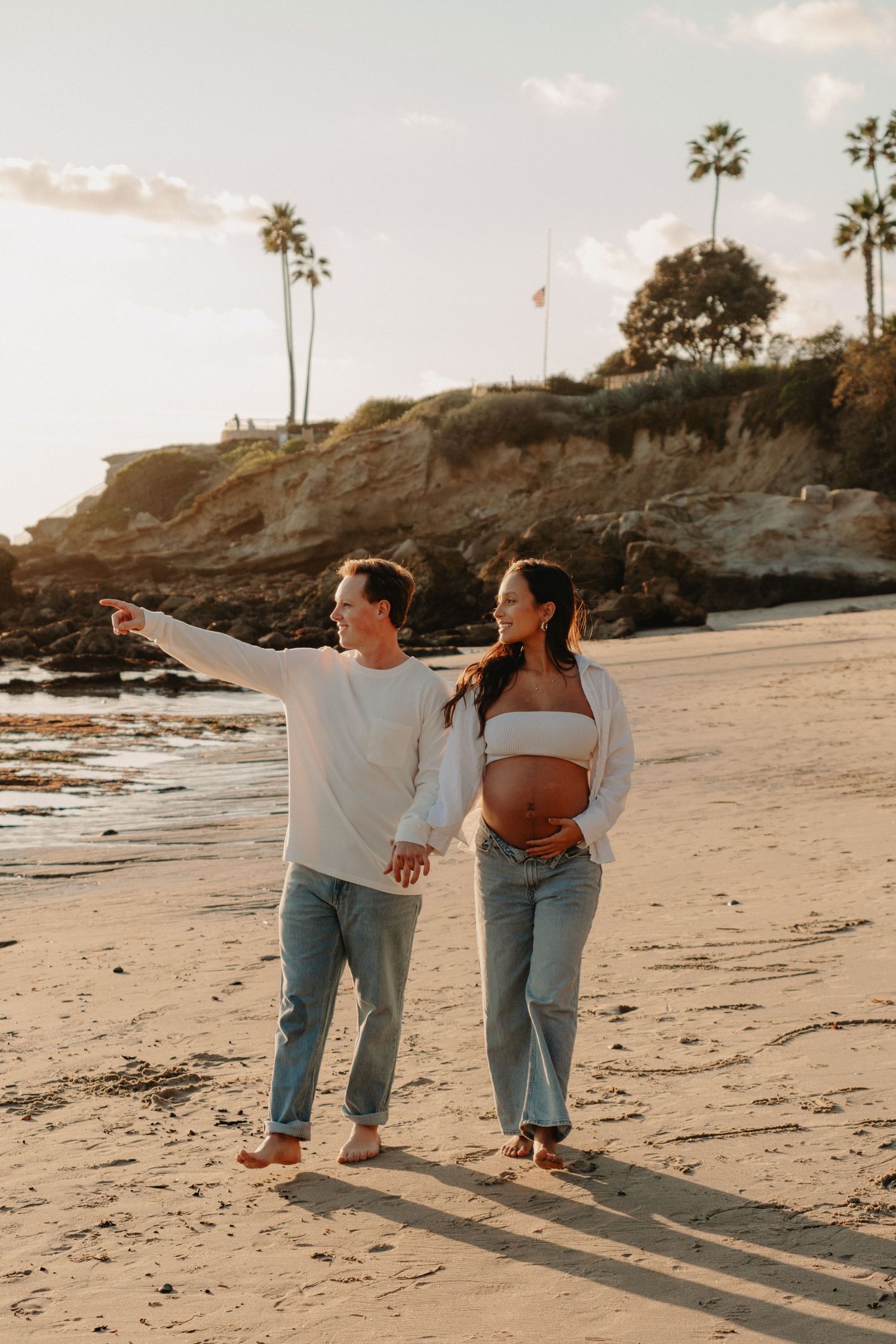 Pregnant couple walking hand-in-hand on a sandy beach at sunset, with palm trees and rocky cliffs in the background.