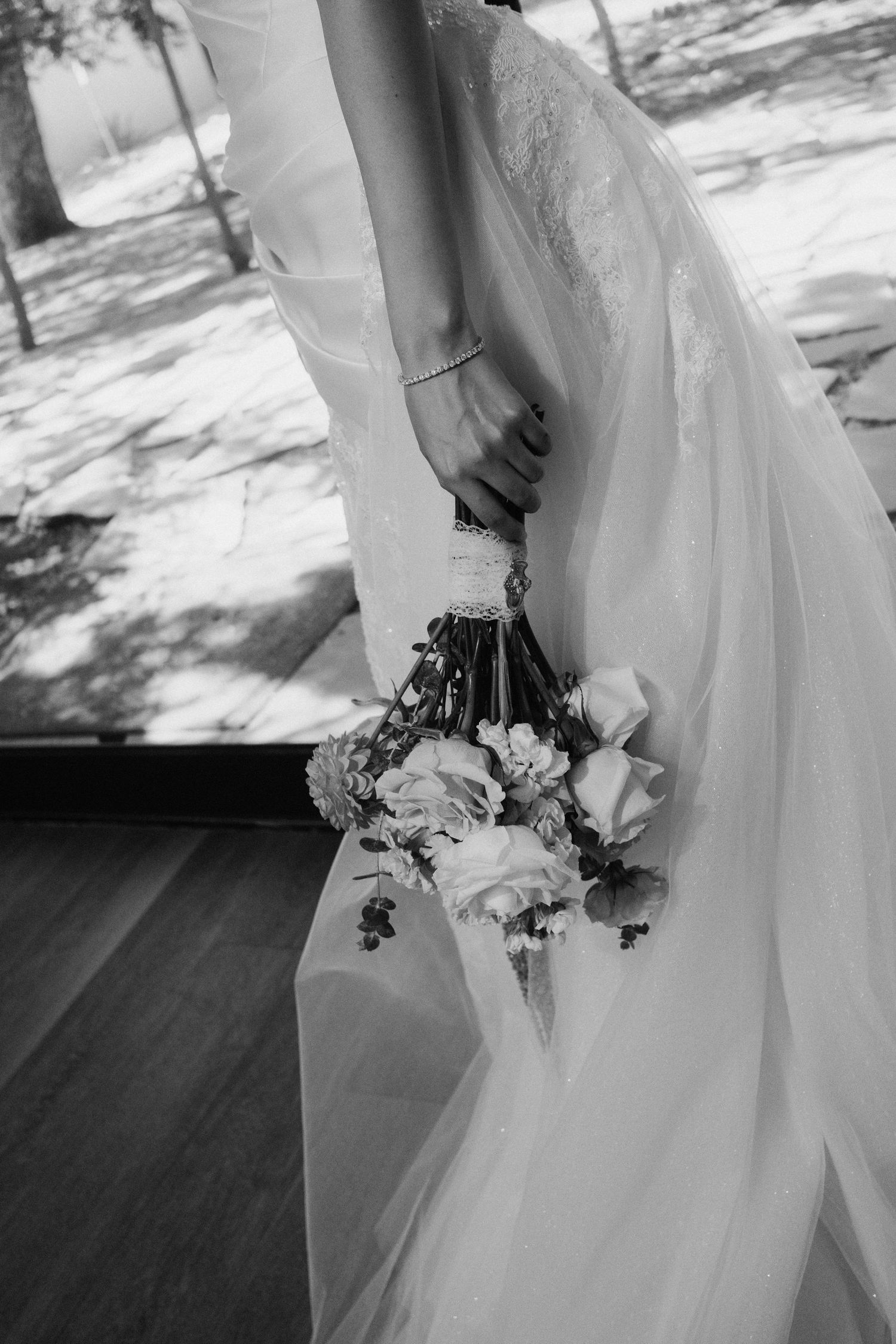 Bride in white lace gown holds elegant floral bouquet with flowing veil in black and white wedding photo.