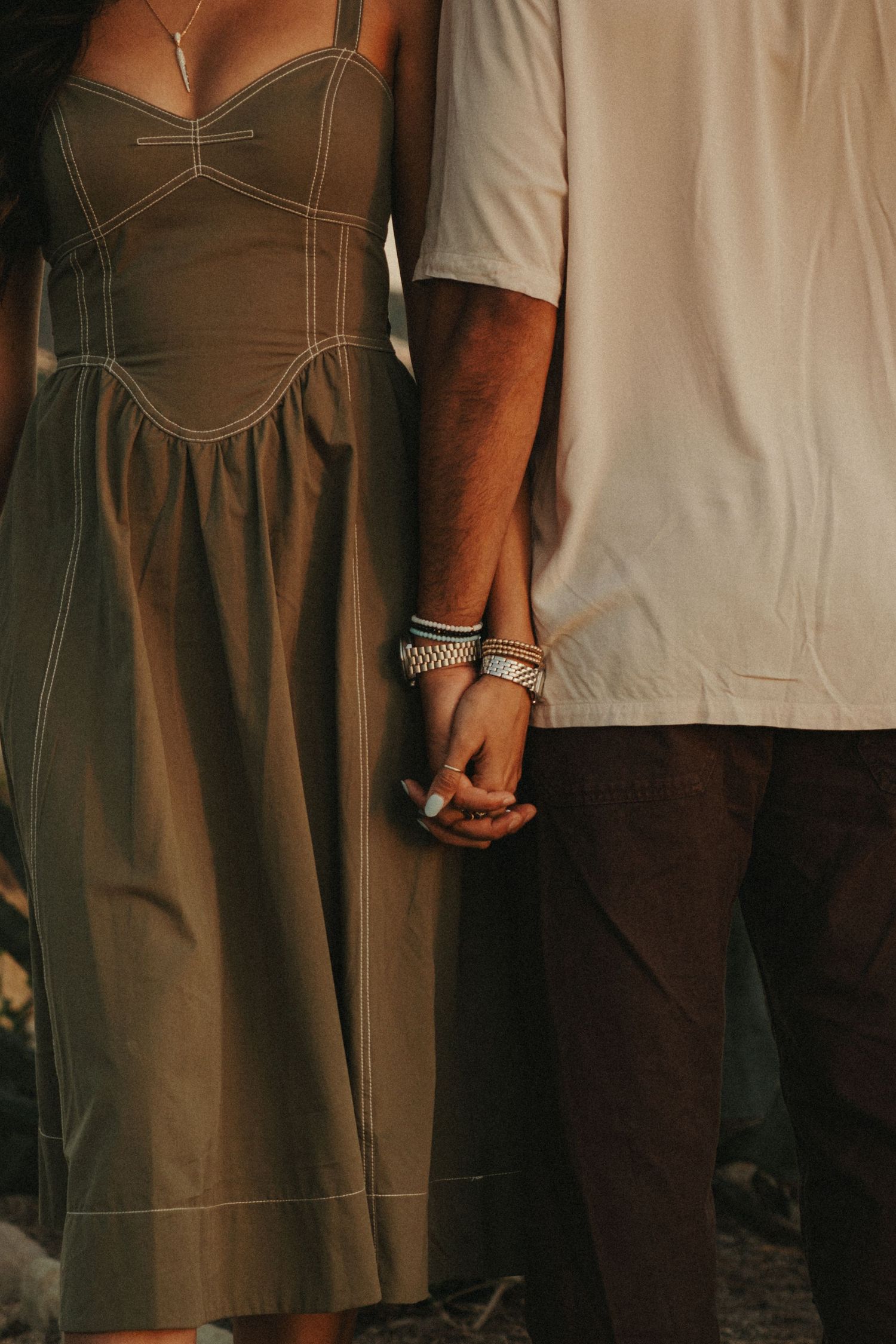 A couple holds hands tenderly, the woman wearing a green corset dress and silver bracelets in warm light.