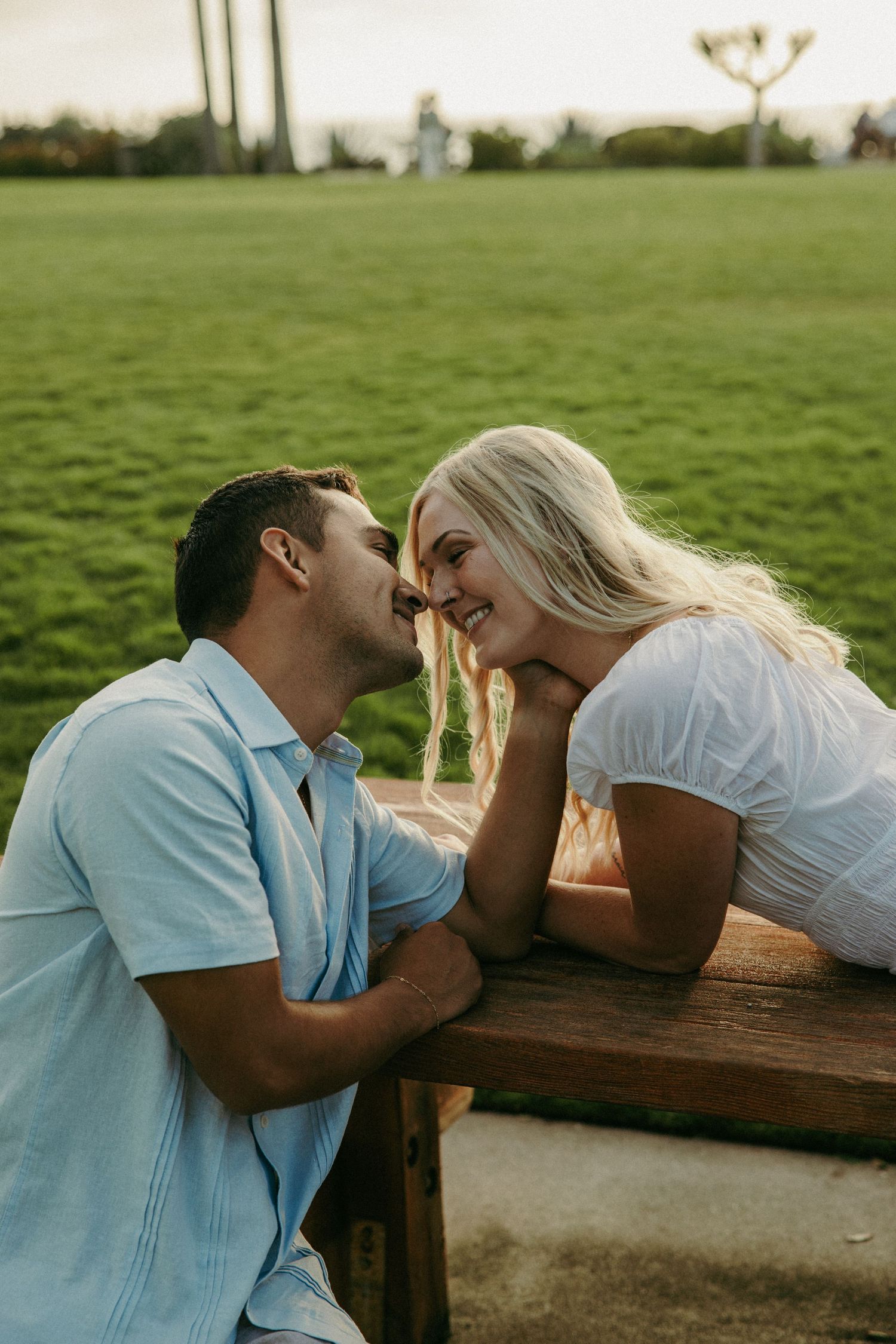 A couple shares a romantic moment at an outdoor wooden table, with a lush green field in the background.