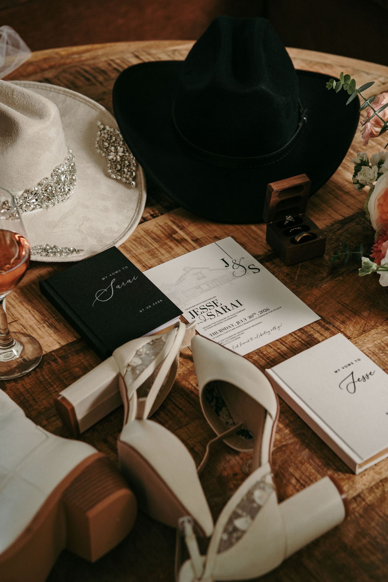 Rustic wedding flat lay with cowboy hats, white heels, invitations, vow books, and ring box on a wooden table.