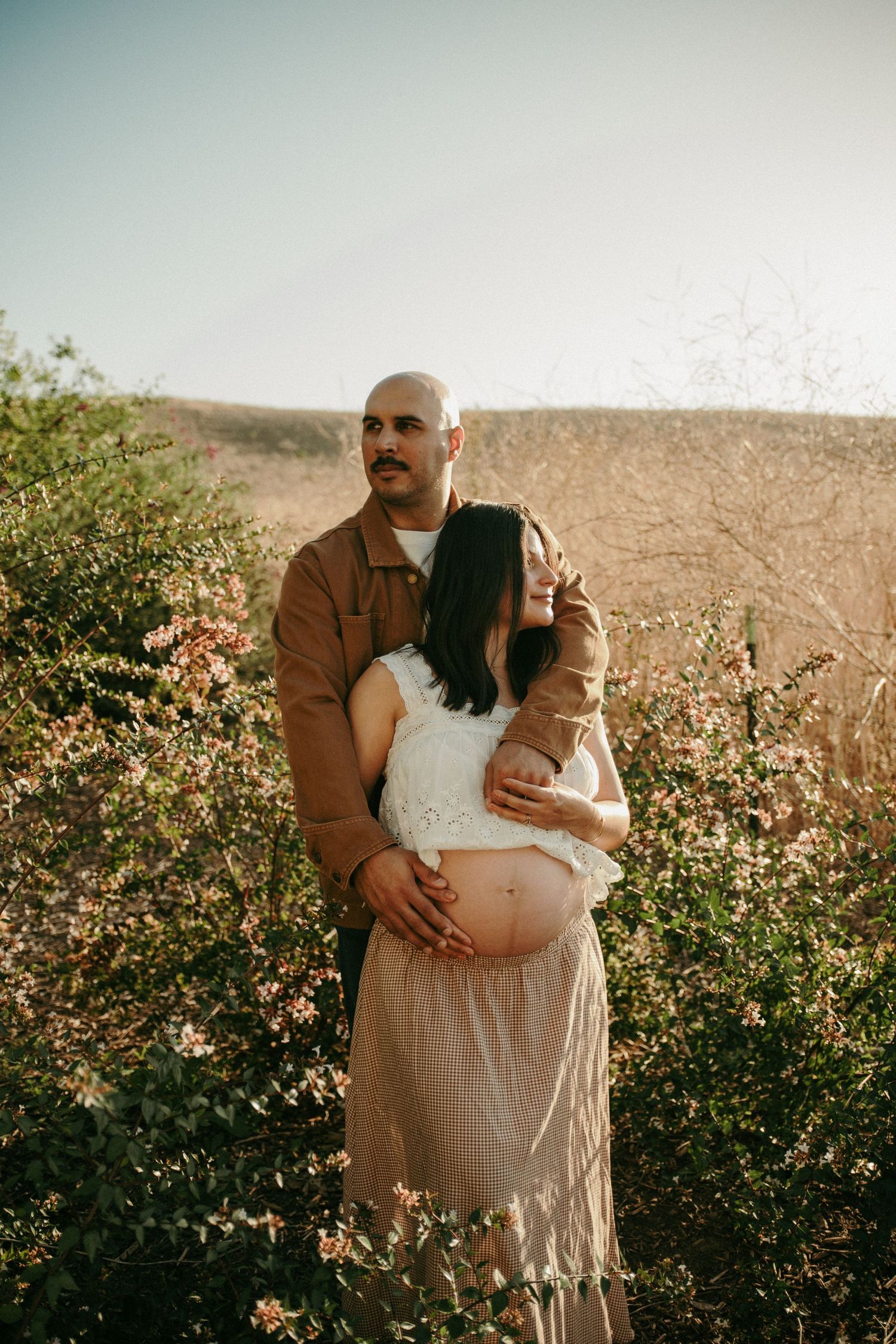 Couple embracing outdoors, pregnant woman in white crop top and beige skirt, man in brown jacket, surrounded by blooming bushes.