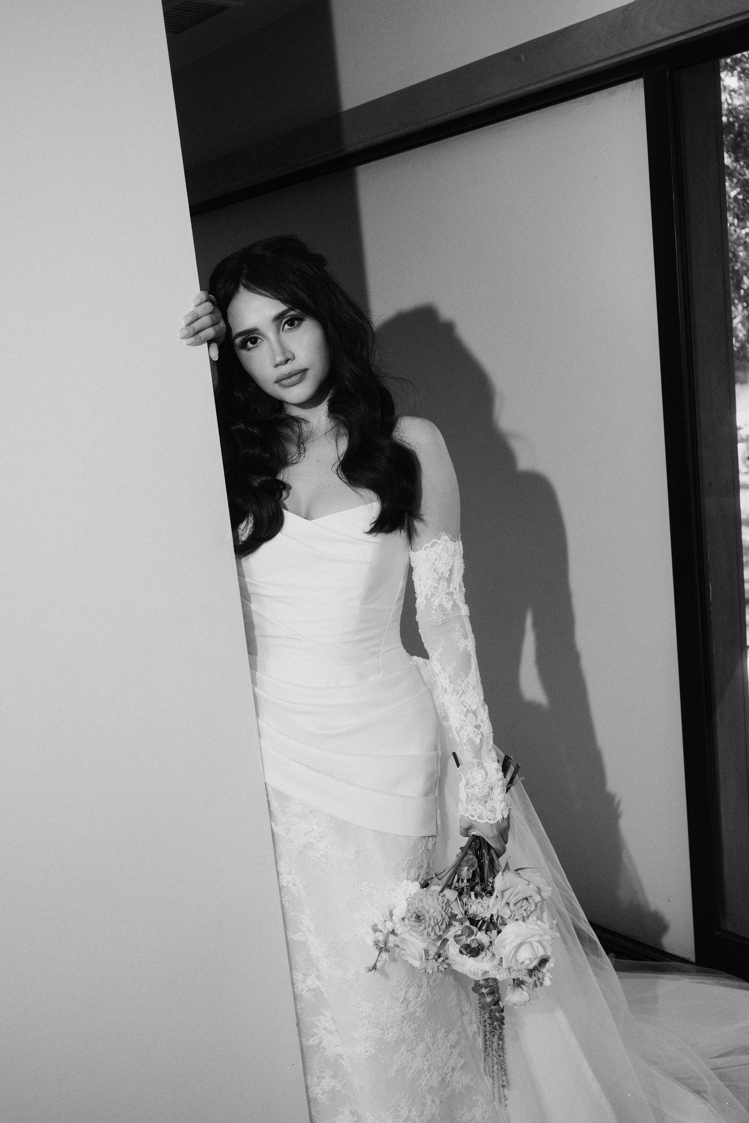 Elegant bride in a fitted lace wedding gown holding a bouquet, posing gracefully in black and white photo.